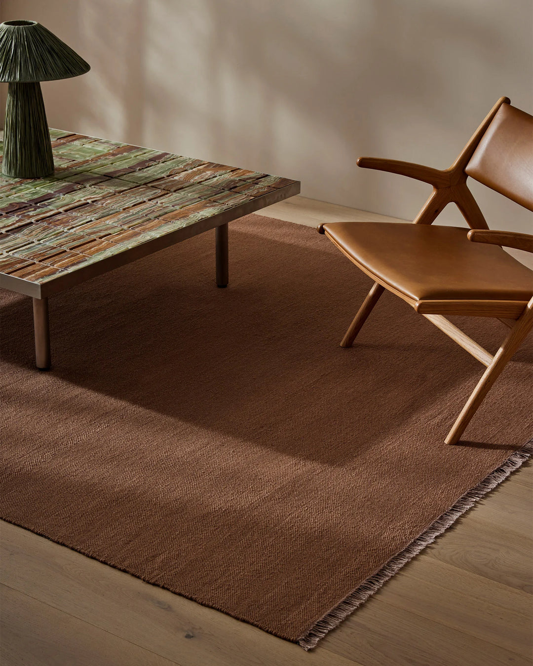 Mid-century modern living space featuring a rich brown wool rug with fringed edge, paired with cognac leather seating and sculptural timber furniture in warm neutral tones