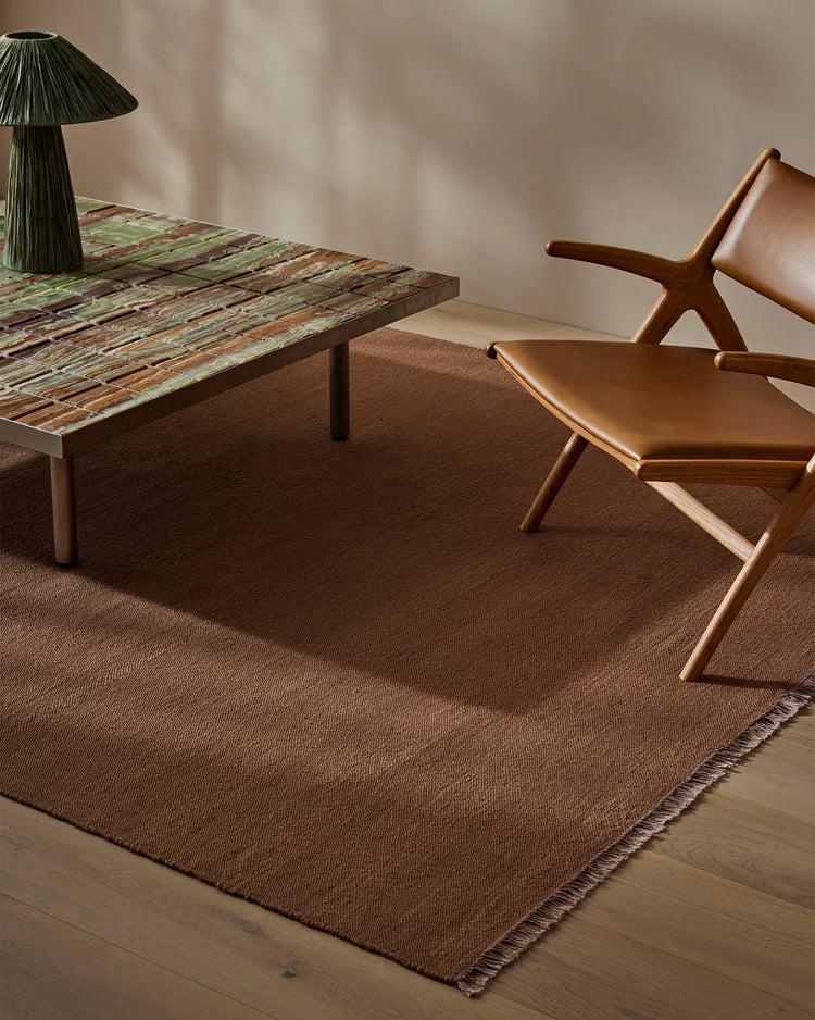 Mid-century modern living space featuring a rich brown wool rug with fringed edge, paired with cognac leather seating and sculptural timber furniture in warm neutral tones