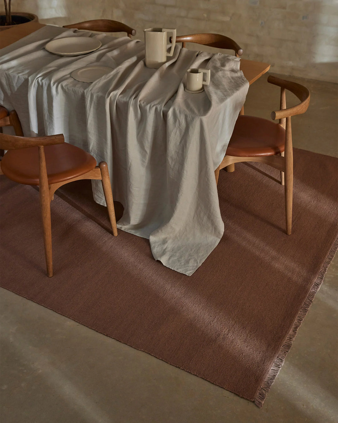 Mid-century dining setting with bone linen tablecloth draped over round table, timber chairs with cognac leather seats, and rosewood wool rug anchoring the scene against neutral concrete backdrop