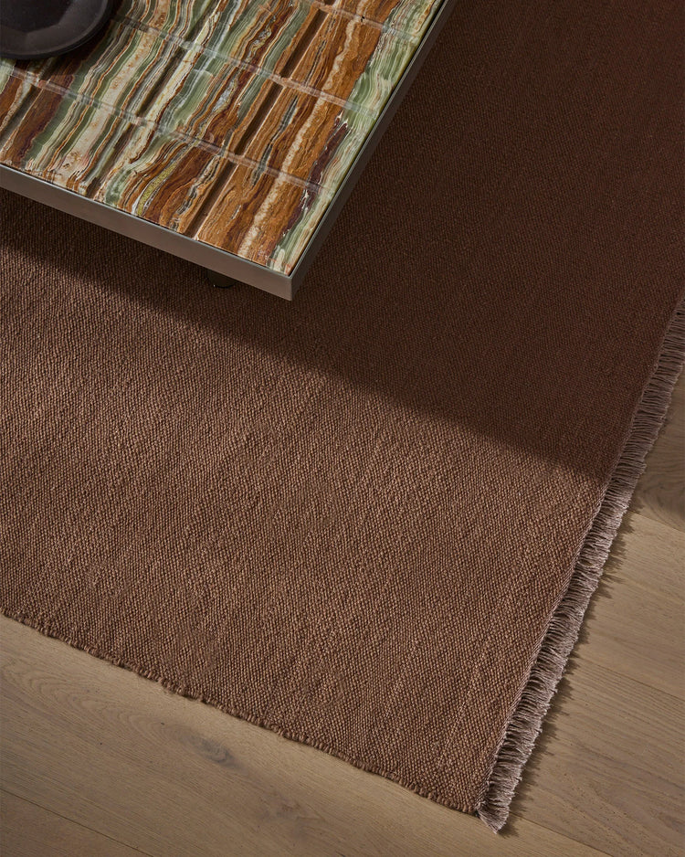 Earthy, layered interior with deep mauve wool rug on light timber flooring, styled with striped earth-tone artwork in ochre, sage and terracotta tones