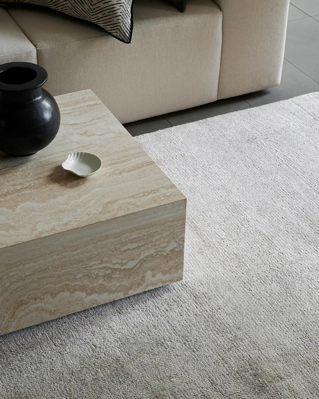 Neutral living room with cream linen sofa, travertine coffee table, and Almonte Oyster low-traffic rug in soft greyscale tones layered with black ceramic accent pieces