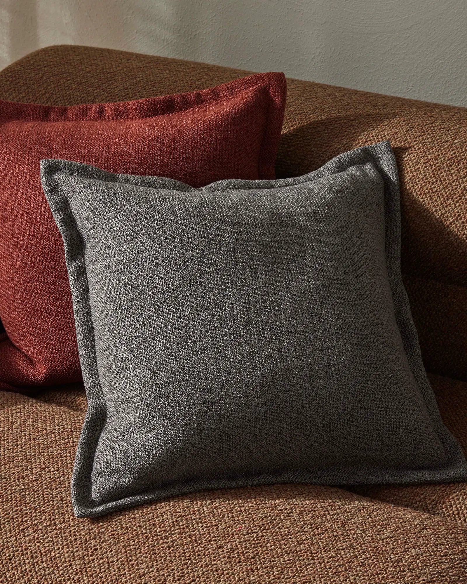 Charcoal and burgundy cushions layered on natural textured lounge chair, warm earth tones and soft linen fabrics create cosy minimalist living room styling
