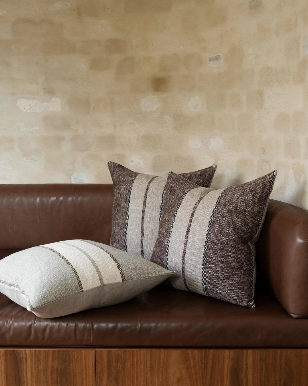 Striped linen-blend cushions in earthy taupe and cream tones arranged on a cognac leather sofa against weathered plaster wall, mid-century modern styling