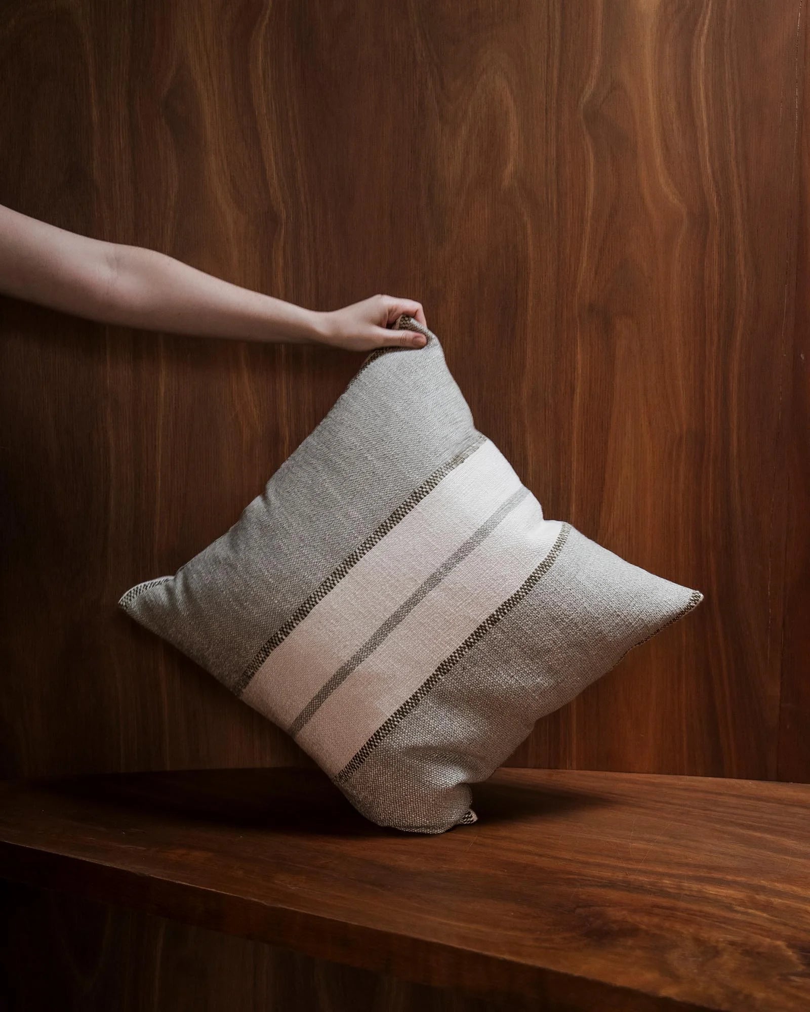 Neutral linen blend cushion with tonal stripe detail held against rich walnut wood paneling, showcasing warm minimalist interior styling