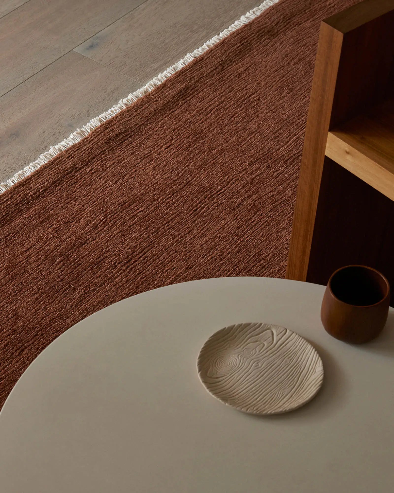 Warm terracotta wool rug with fringe edge anchors minimalist dining scene, pairing earthy tones with natural wood and cream ceramics