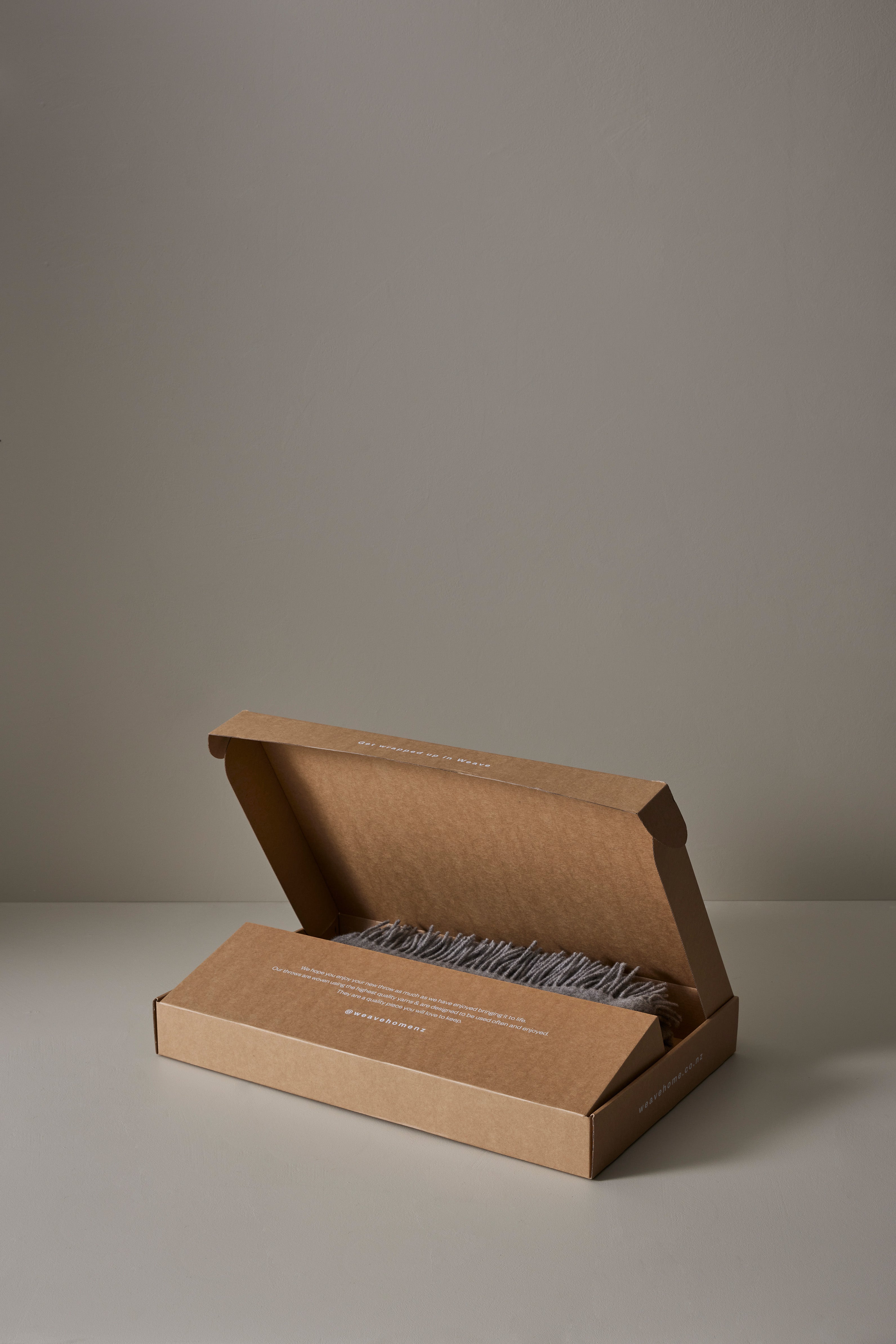 Kraft cardboard gift box open to reveal a folded wool throw with fringe detail, neutral taupe and grey palette against soft greyscale backdrop, sustainable packaging styling