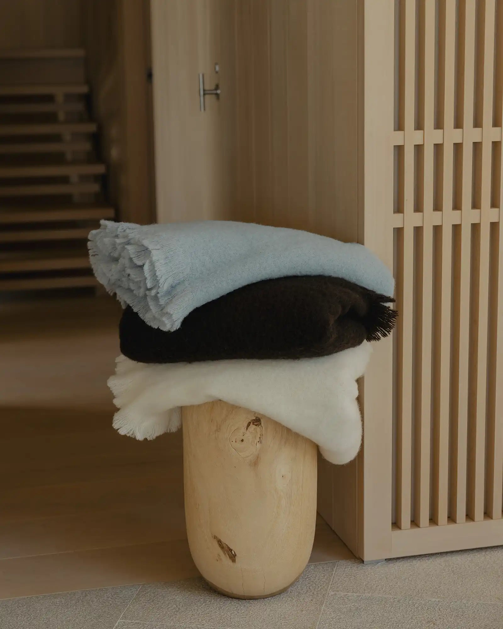 Neutral entryway styled with stacked wool blend throws in cream, charcoal and sage grey draped over timber post, natural wood flooring and timber slat wall panelling
