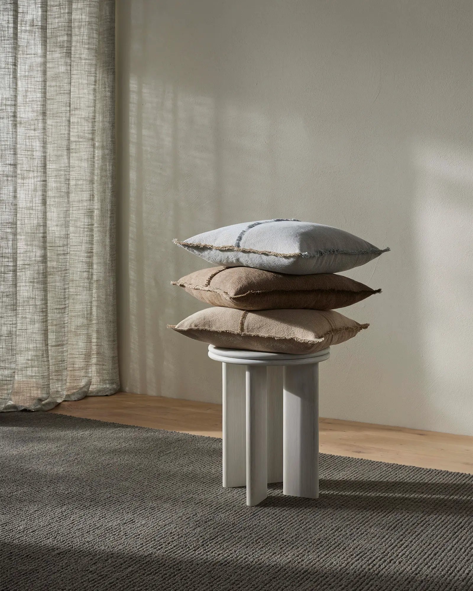 Stacked linen cushions in soft neutrals—haze, cashmere, and cocoa—on a white pedestal amid natural light, textured rug, and oatmeal linen curtains
