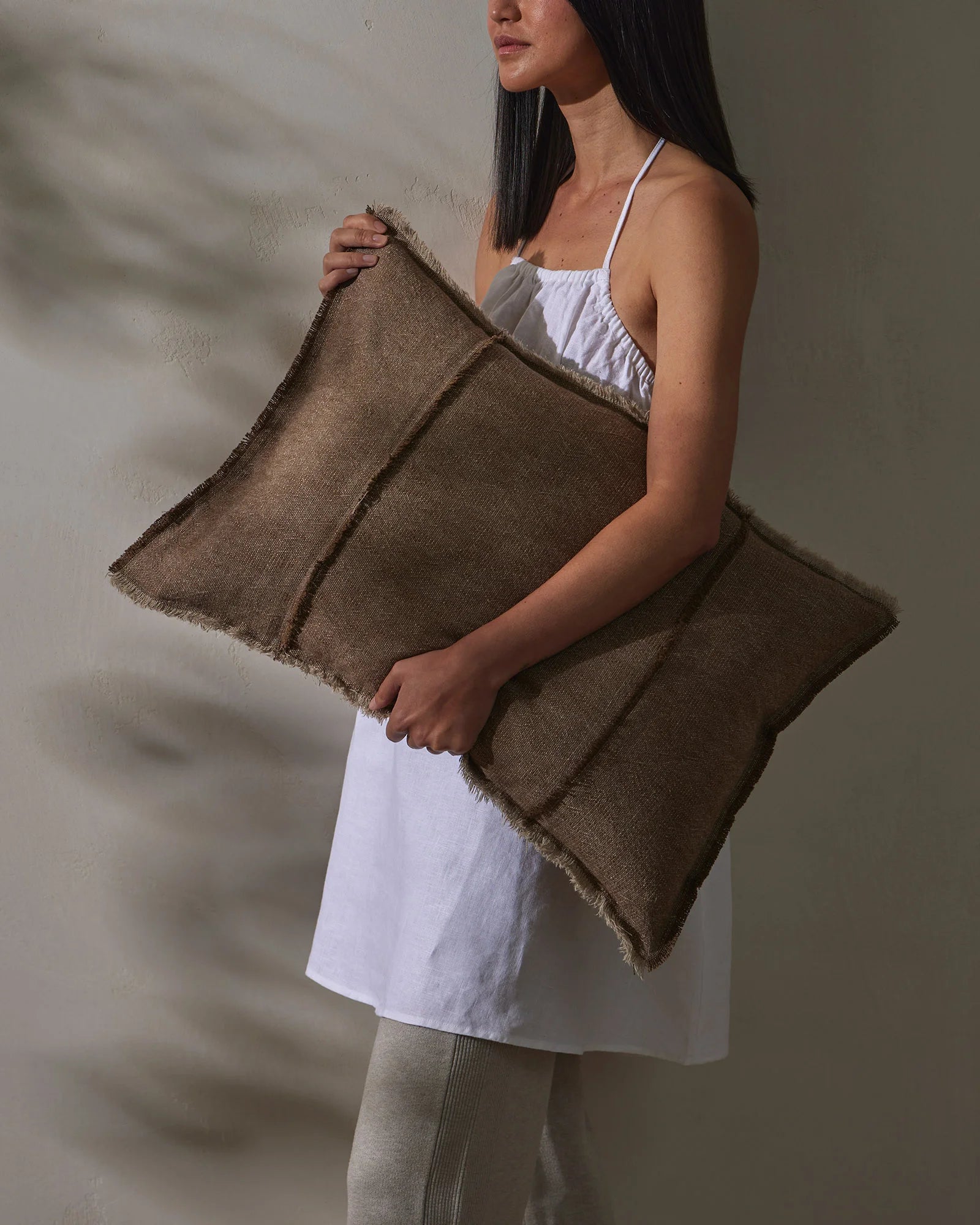 Woman holding taupe linen cushions against white textured wall, styled with white cotton dress and neutral palette, showcasing soft woven textures and earthy tones