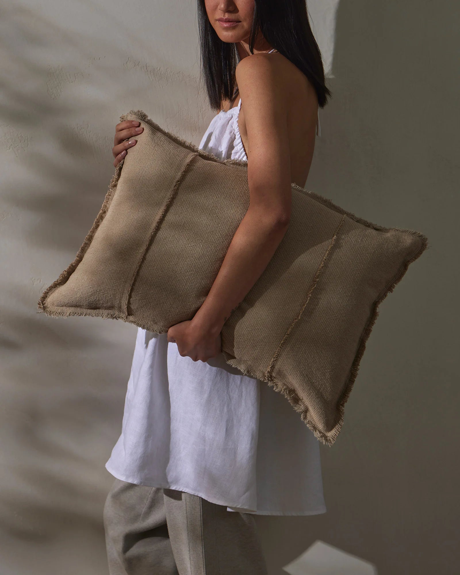 Woman in white linen dress holding oversized natural linen cushion with raw frayed edges against textured white wall, minimalist neutral aesthetic