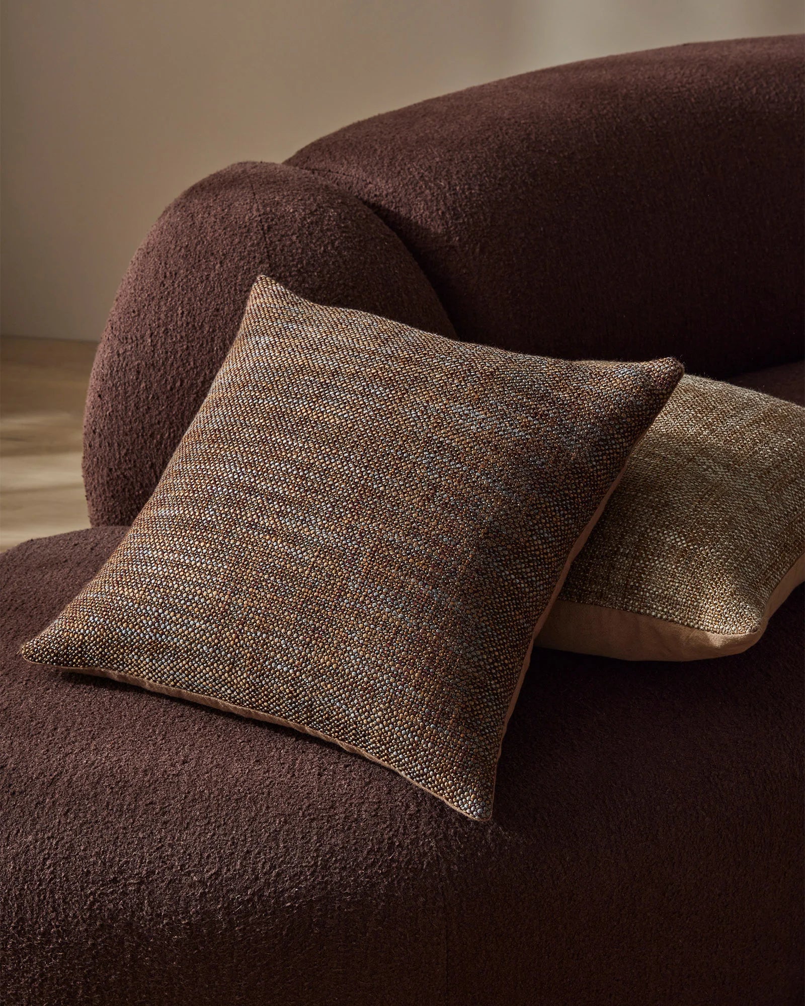 Earthy taupe and chocolate cushions with textured linen-look weave arranged on a burgundy upholstered chair in warm neutral styling