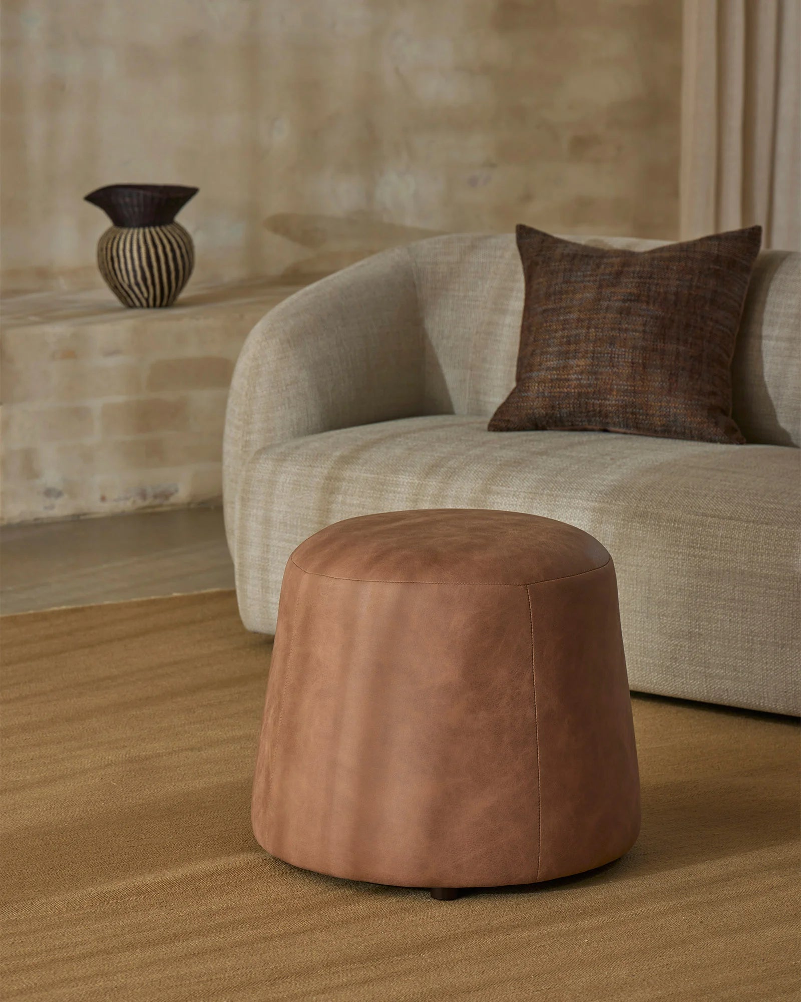 Neutral minimalist living room with cream curved armchair, warm tan wool rug, chocolate linen cushion and cognac leather ottoman on neutral textured backdrop