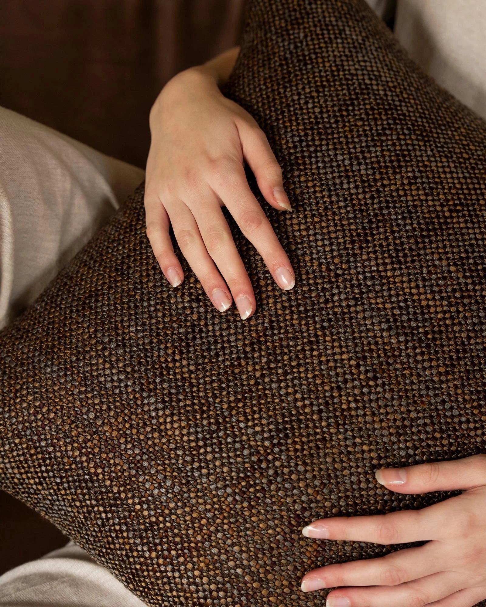 Hands touching textured brown cushion showing woven weave detail, warm neutral tones with layered earth palette and soft linen styling