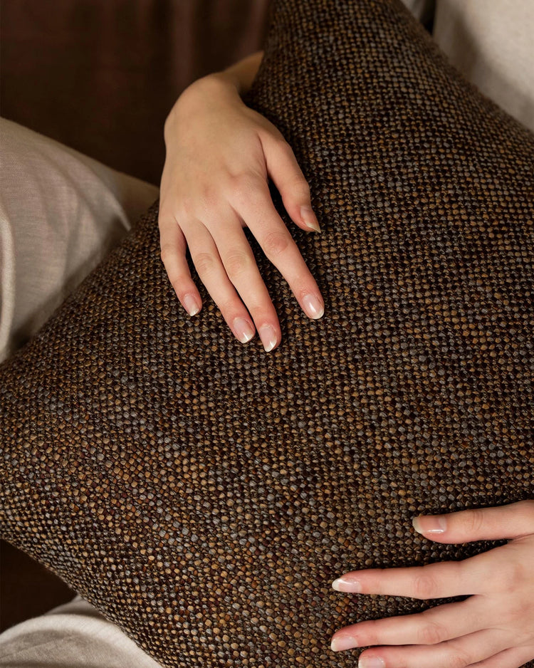 Hands touching textured brown cushion showing woven weave detail, warm neutral tones with layered earth palette and soft linen styling