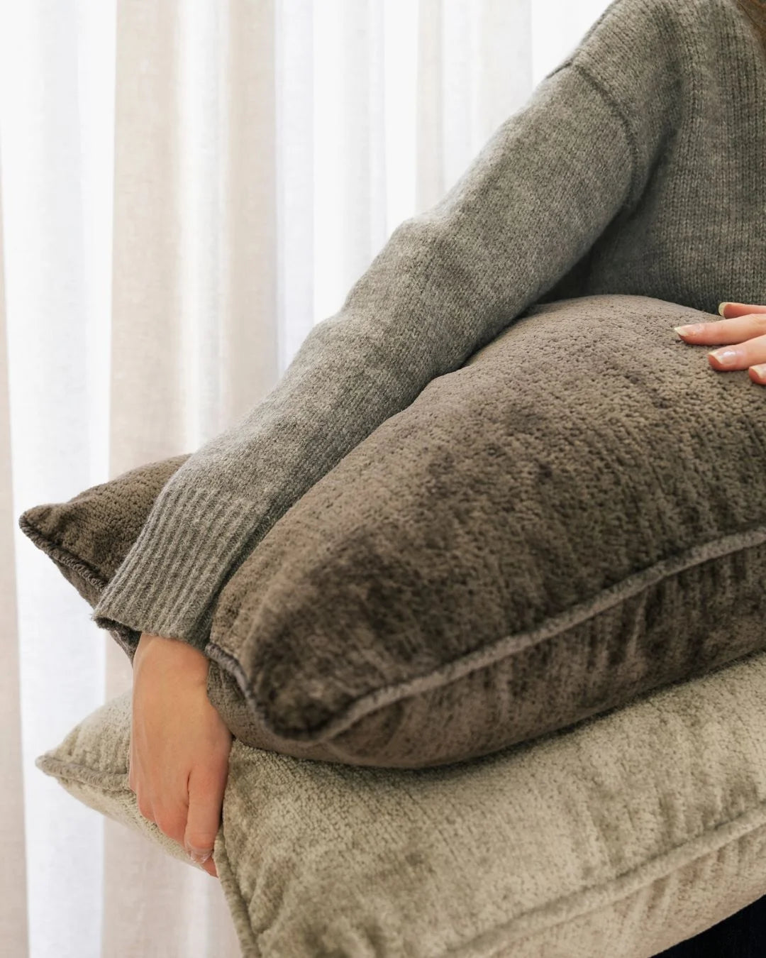 Neutral toned lifestyle shot of stacked Tempo cushions in soft grey and beige textures layered with ribbed knitwear against striped linen curtains