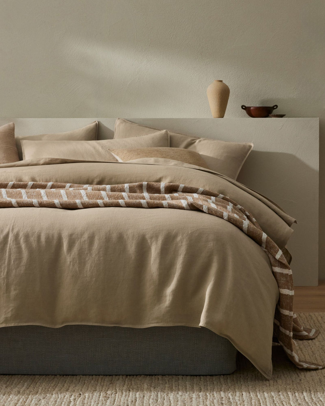 Minimalist bedroom with neutral linen bedding, layered cream and taupe tones, natural geometric patterned throw across bed, soft shadows on white headboard, warm wood accents