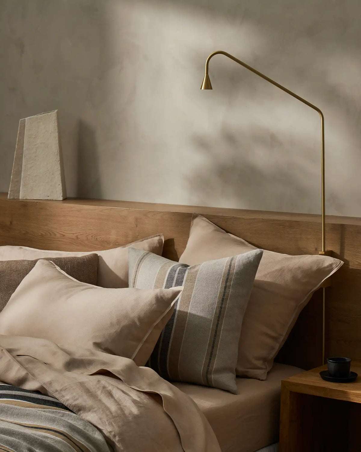 Neutral bedroom with wooden bed frame, layered linen bedding in cream and taupe tones, striped cushion, soft natural light casting shadows on pale walls
