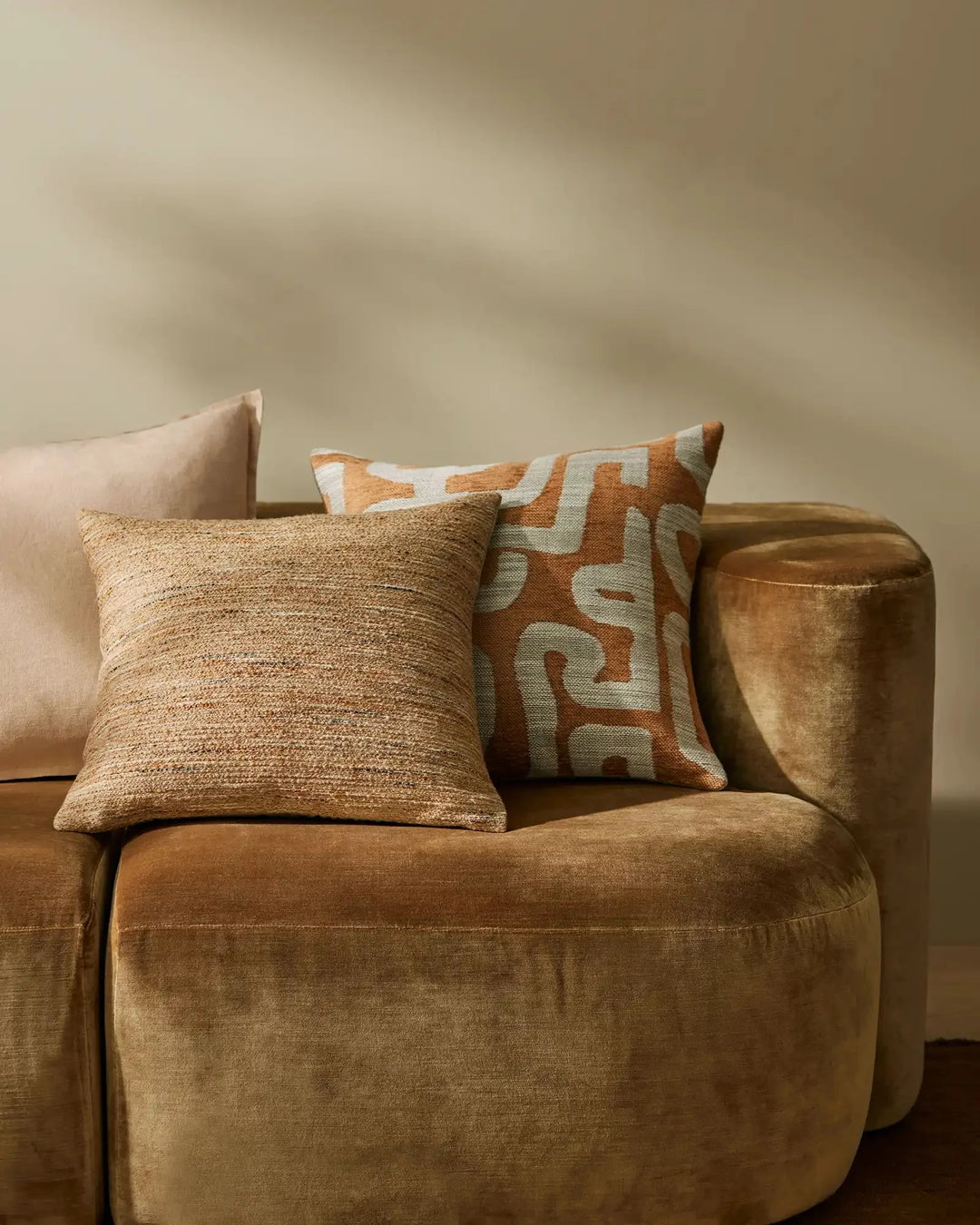 Warm terracotta and cream cushions layered on a camel-toned armchair against a soft neutral backdrop, mixing textured linens with geometric patterns for a contemporary comfort aesthetic