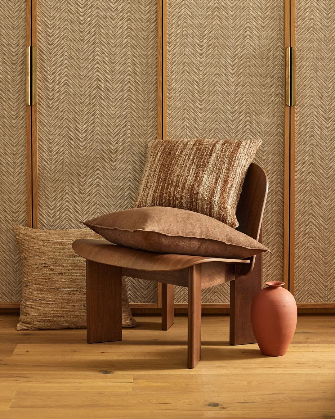Earthy minimalist styling with Gigi, Vista and Fiore cushions layered on wooden chair against herringbone wall panelling in warm terracotta and neutral tones