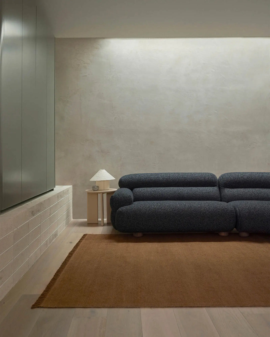 Minimalist living room with charcoal sofa, warm toffee linen rug, pale timber accents and raw concrete wall for a sophisticated, understated aesthetic