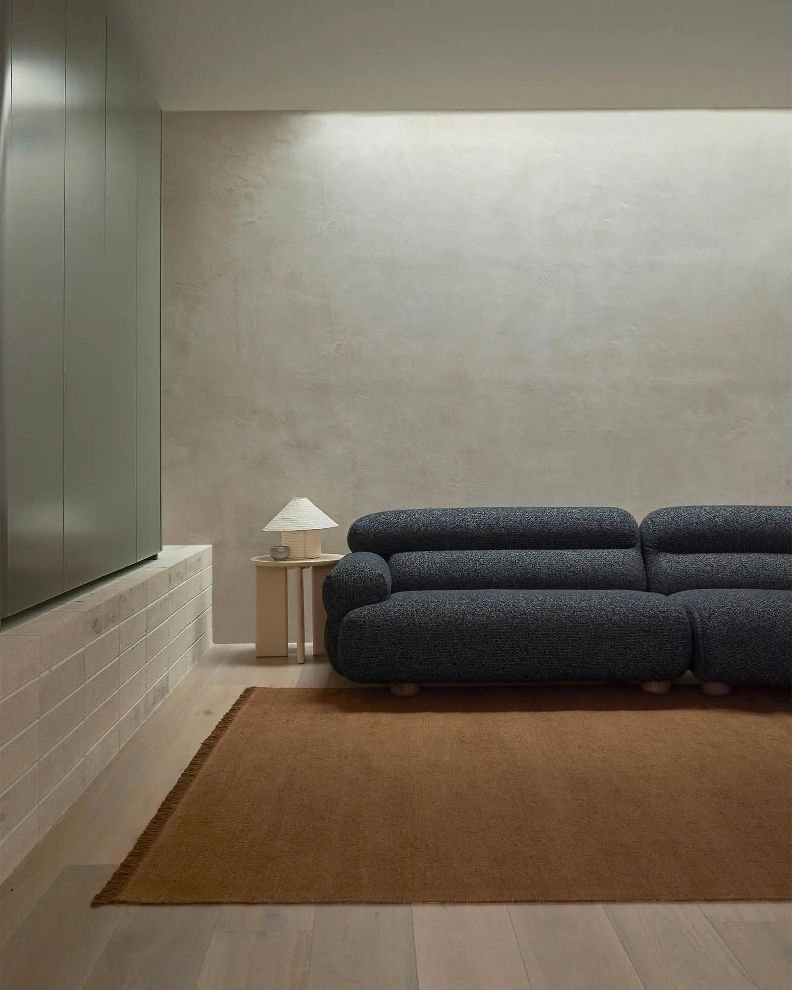 Minimalist living room with charcoal sofa, warm toffee linen rug, pale timber accents and raw concrete wall for a sophisticated, understated aesthetic