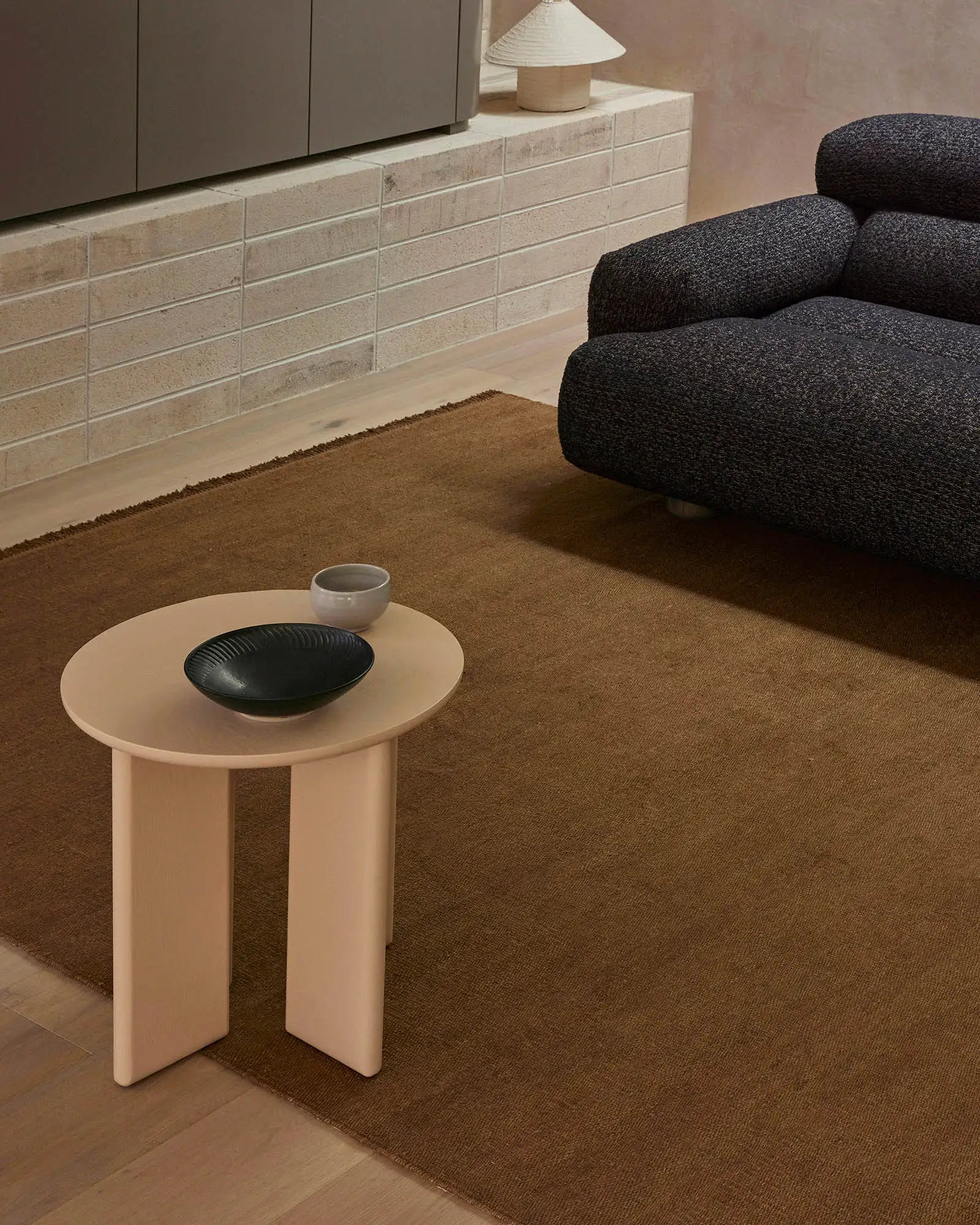 Warm minimalist living space with charcoal curved armchair and cream side table on toffee-toned linen rug, neutral tiled wall backdrop, earthy palette.