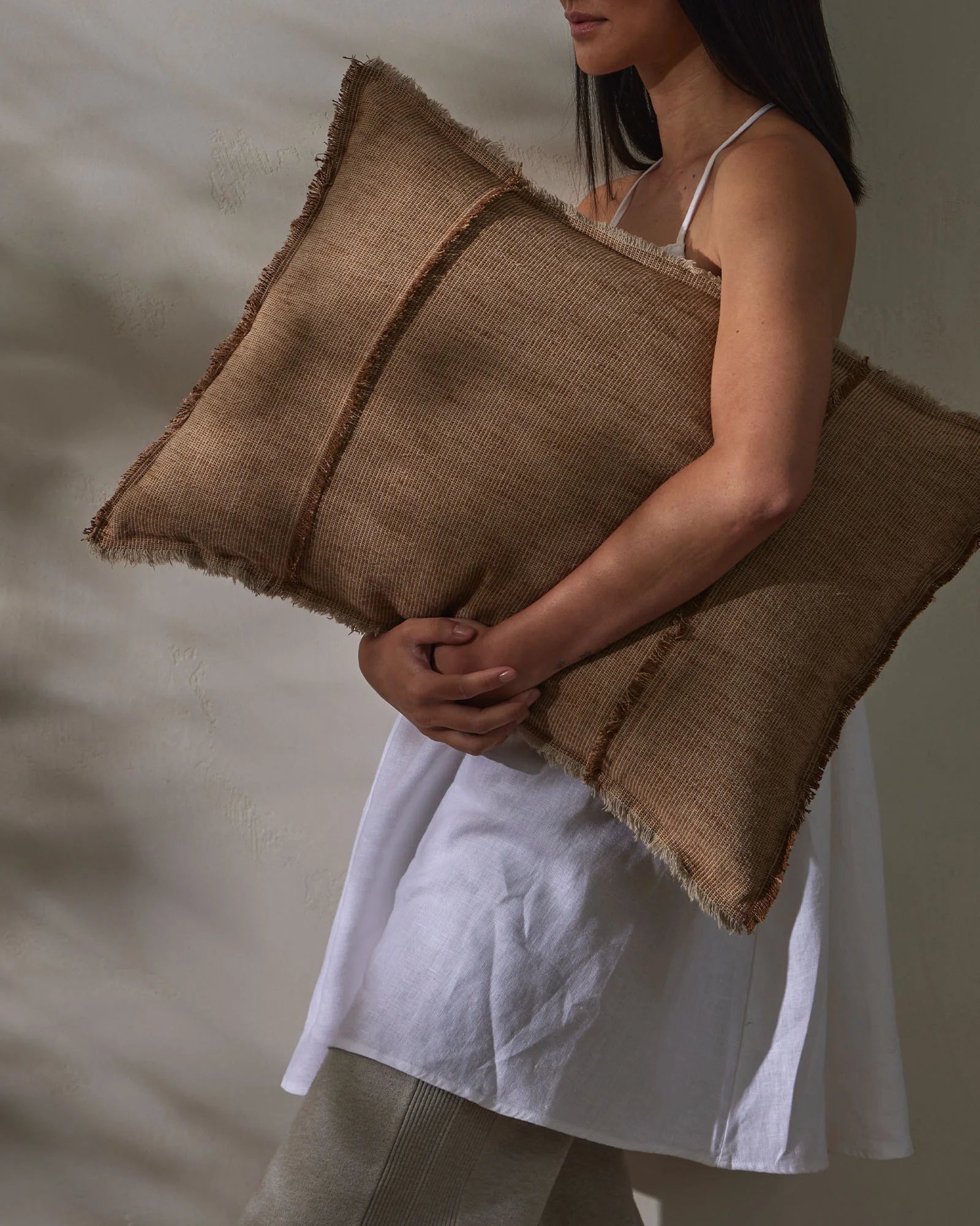 Woman holding layered taupe linen cushions against neutral backdrop, showcasing ribbed texture and raw-edge fringe detail in warm earth tones