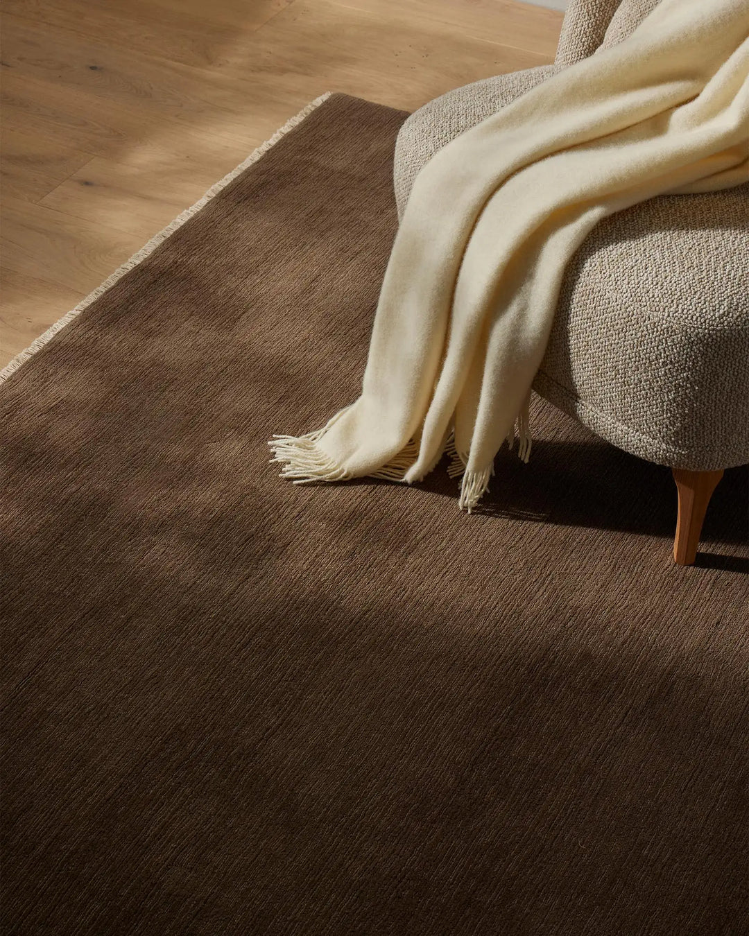 Neutral living room with taupe wool rug, cream throw draped over linen upholstered chair, warm wood flooring and fringe details