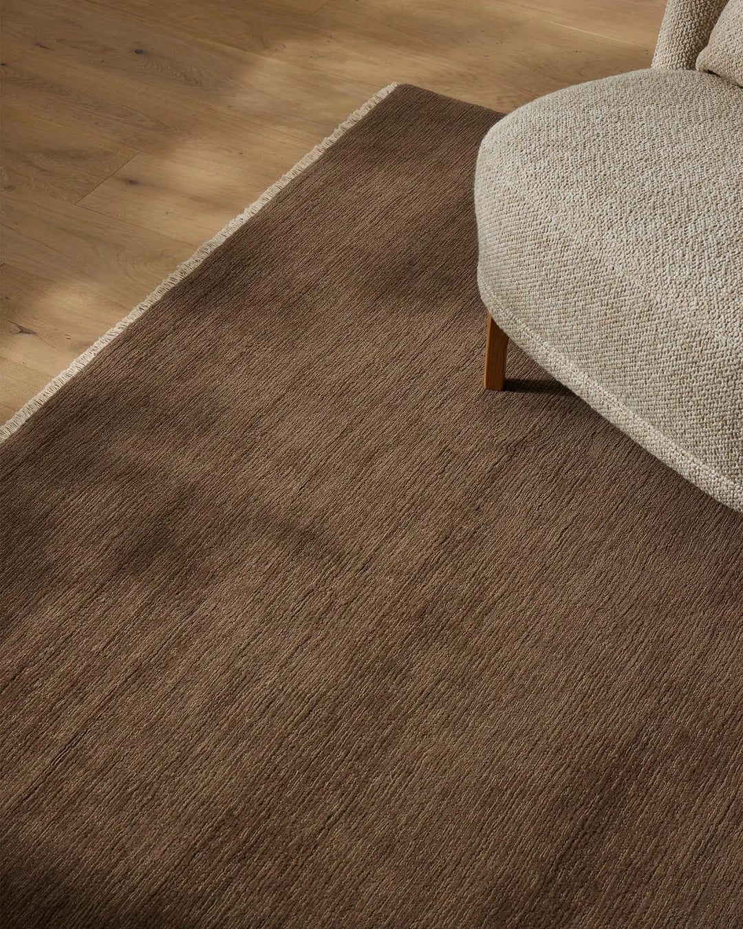 Taupe wool rug with linear texture anchors a warm neutral living space beside a cream upholstered chair and timber flooring