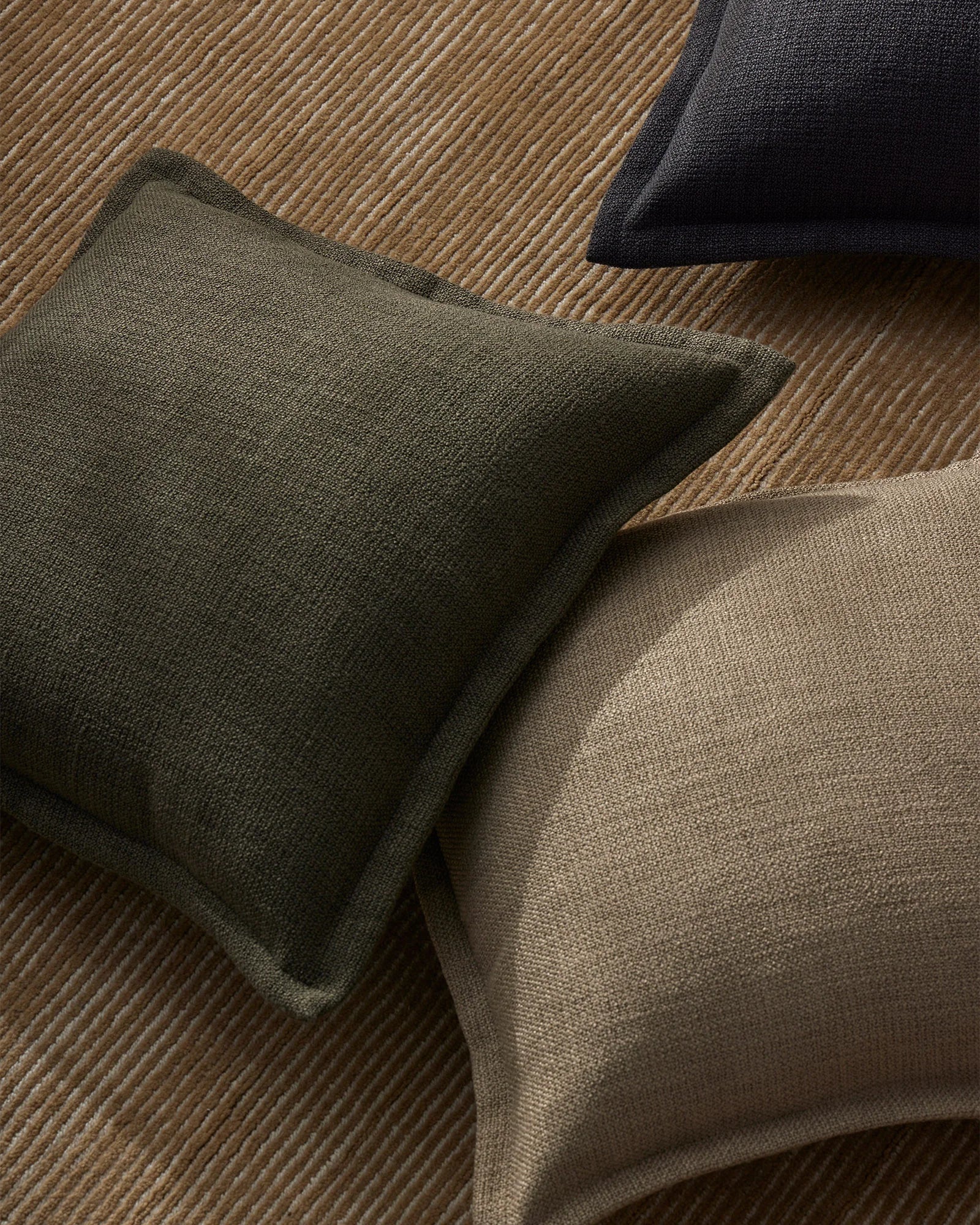 Earthy textured cushions in sage and taupe arranged on a warm wool rug, creating a layered neutral palette with soft tonal contrast