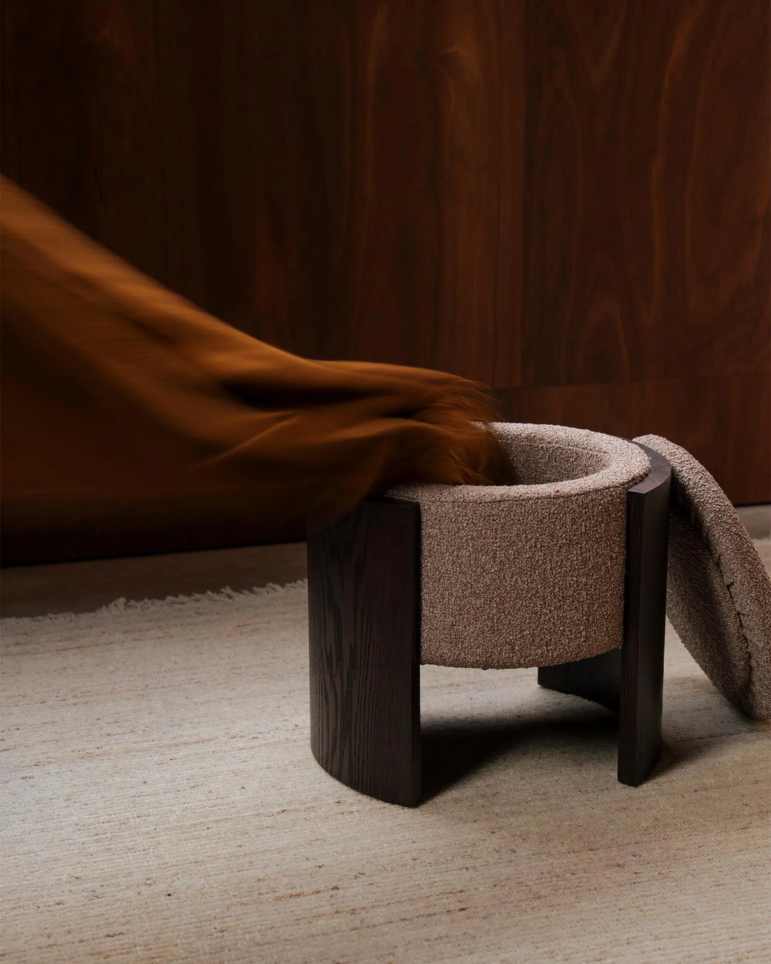 Warm earth-toned lifestyle scene featuring a textured taupe ottoman with dark wood legs, styled with a cinnamon-hued throw against a rich chocolate backdrop and neutral flooring.