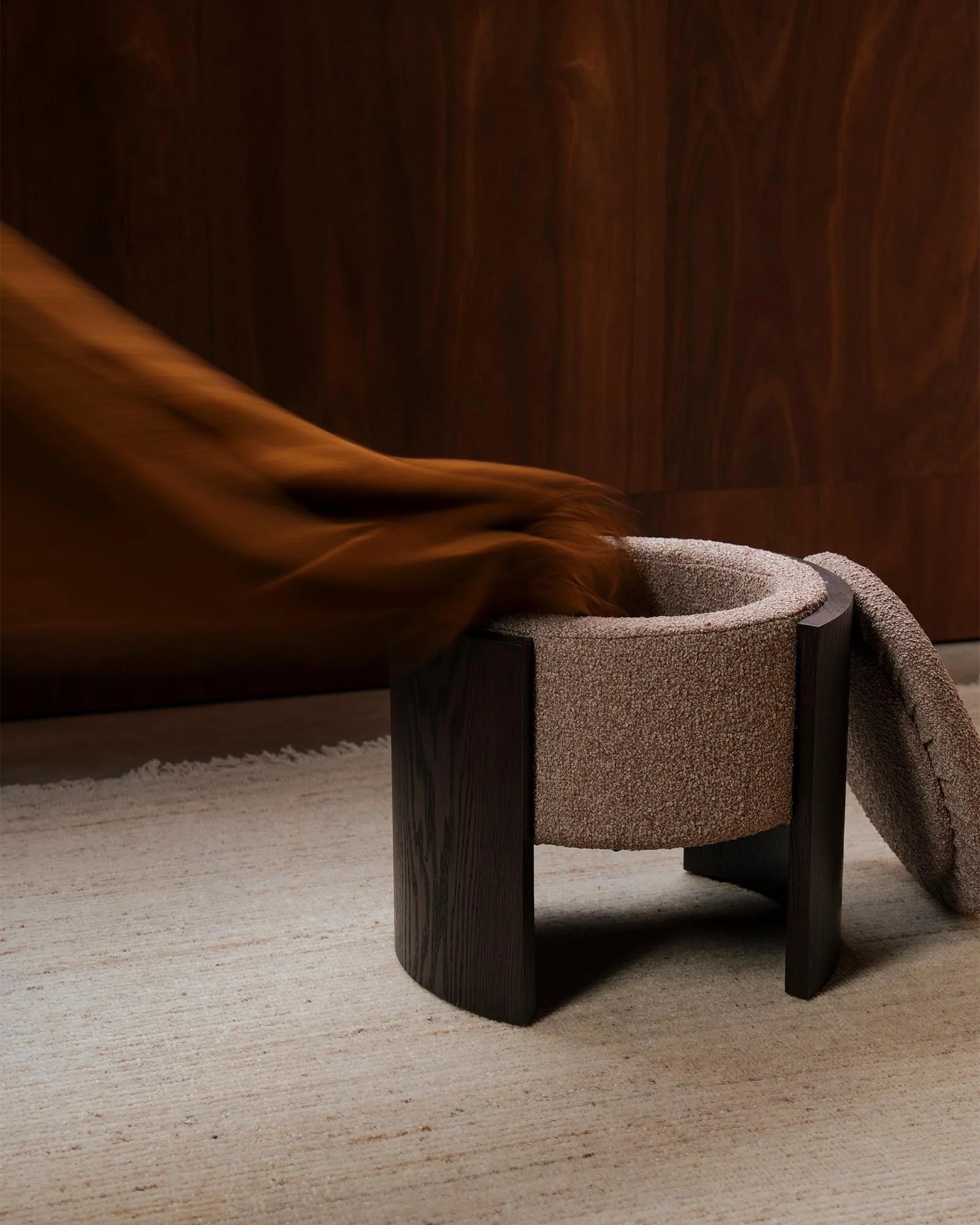 Warm earth-toned lifestyle scene featuring a textured taupe ottoman with dark wood legs, styled with a cinnamon-hued throw against a rich chocolate backdrop and neutral flooring.