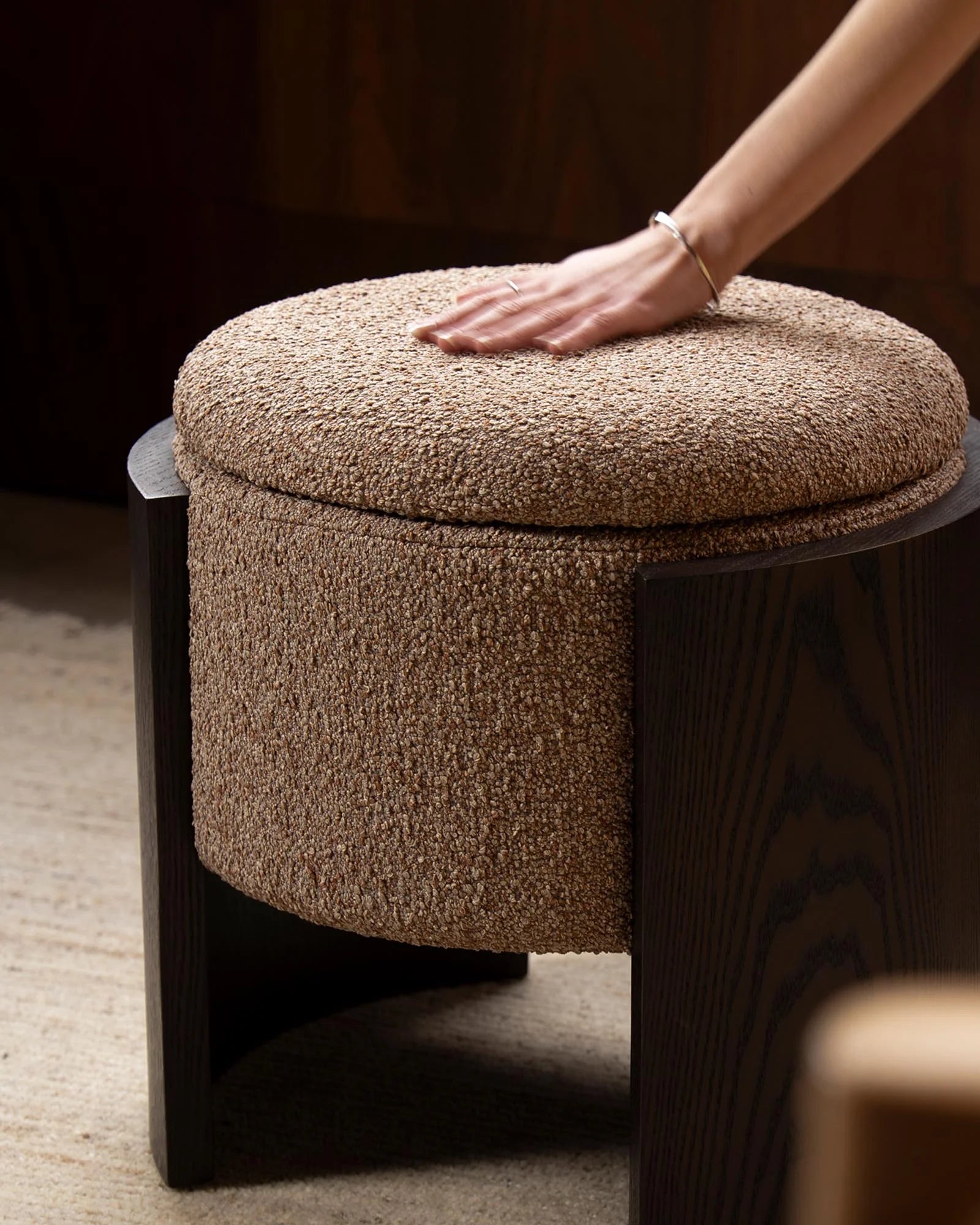 Hand touching textured neutral ottoman with dark wood frame on concrete floor, warm minimalist styling with earthy tones and tactile boucle upholstery