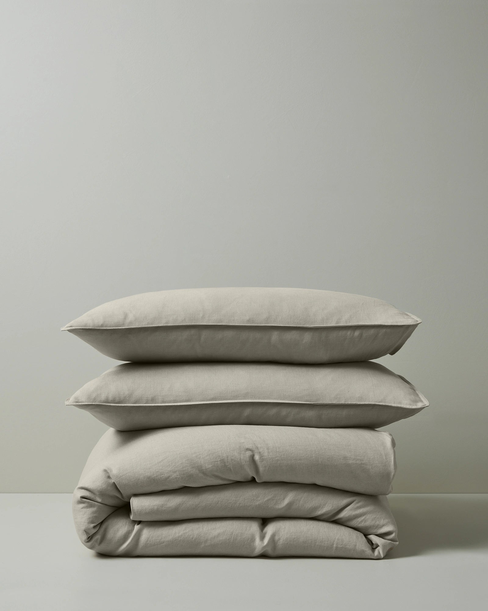 Stacked natural linen bedding in cream and soft sage tones featuring Ravello quilt cover and pillowcase set, showcasing woven texture and relaxed, modern minimalist aesthetic against neutral background