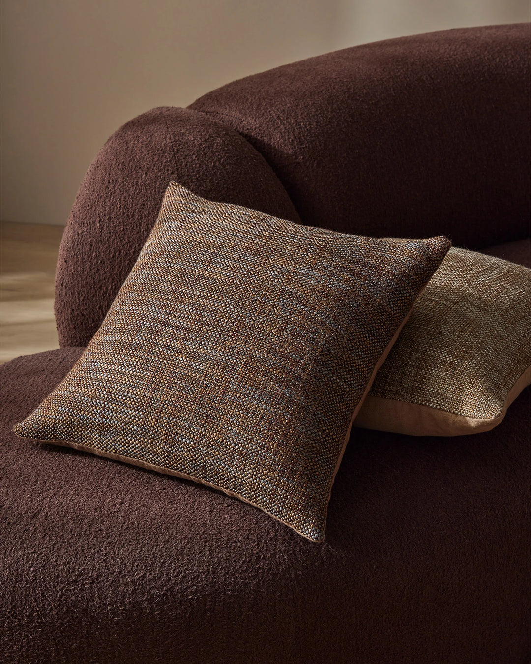 Earthy taupe and chocolate cushions with textured linen-look weave arranged on a burgundy upholstered chair in warm neutral styling