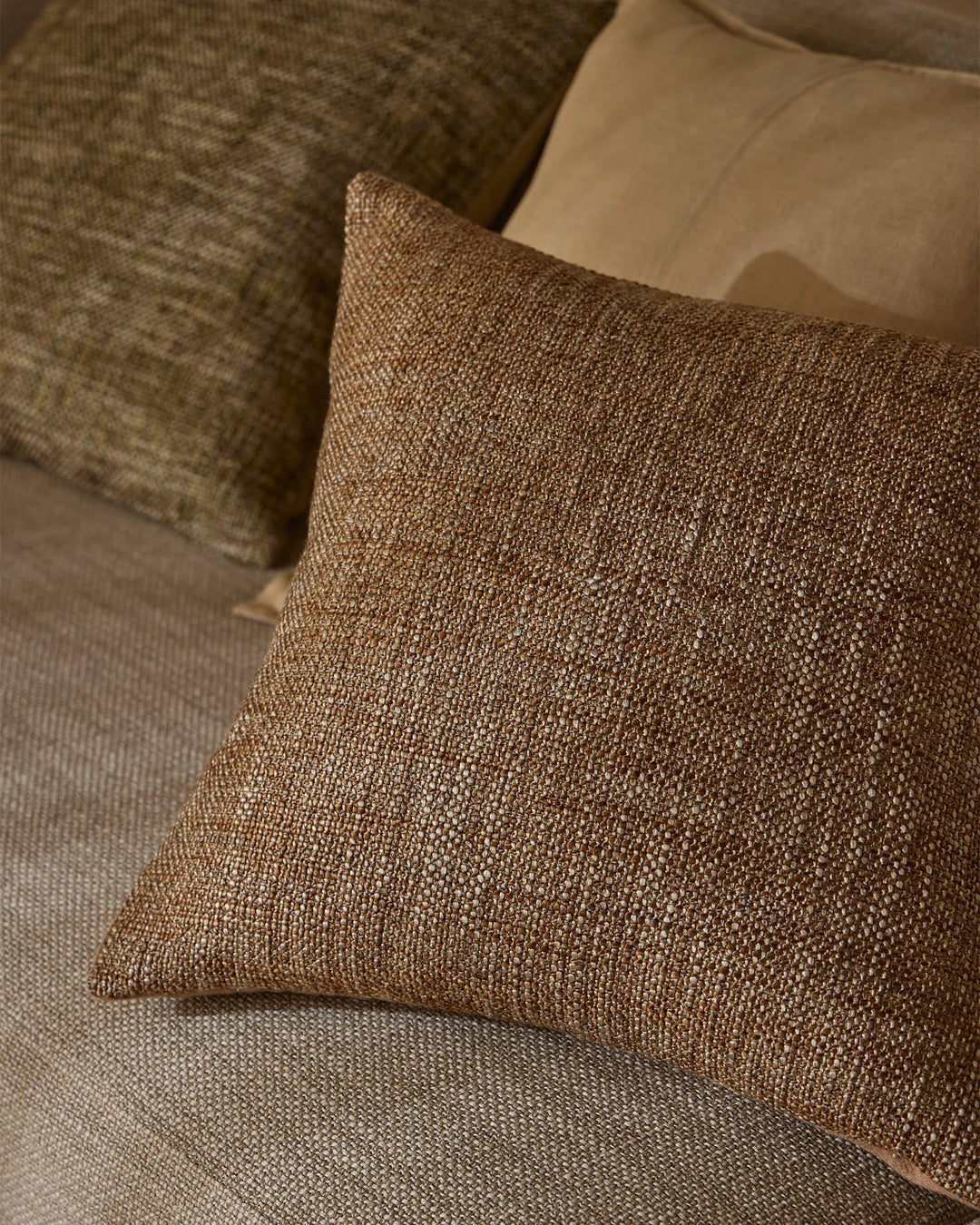 Warm neutral cushions in taupe and toffee tones layered on a cream upholstered sofa, showcasing textured woven fabrics and earthy minimalist interior styling
