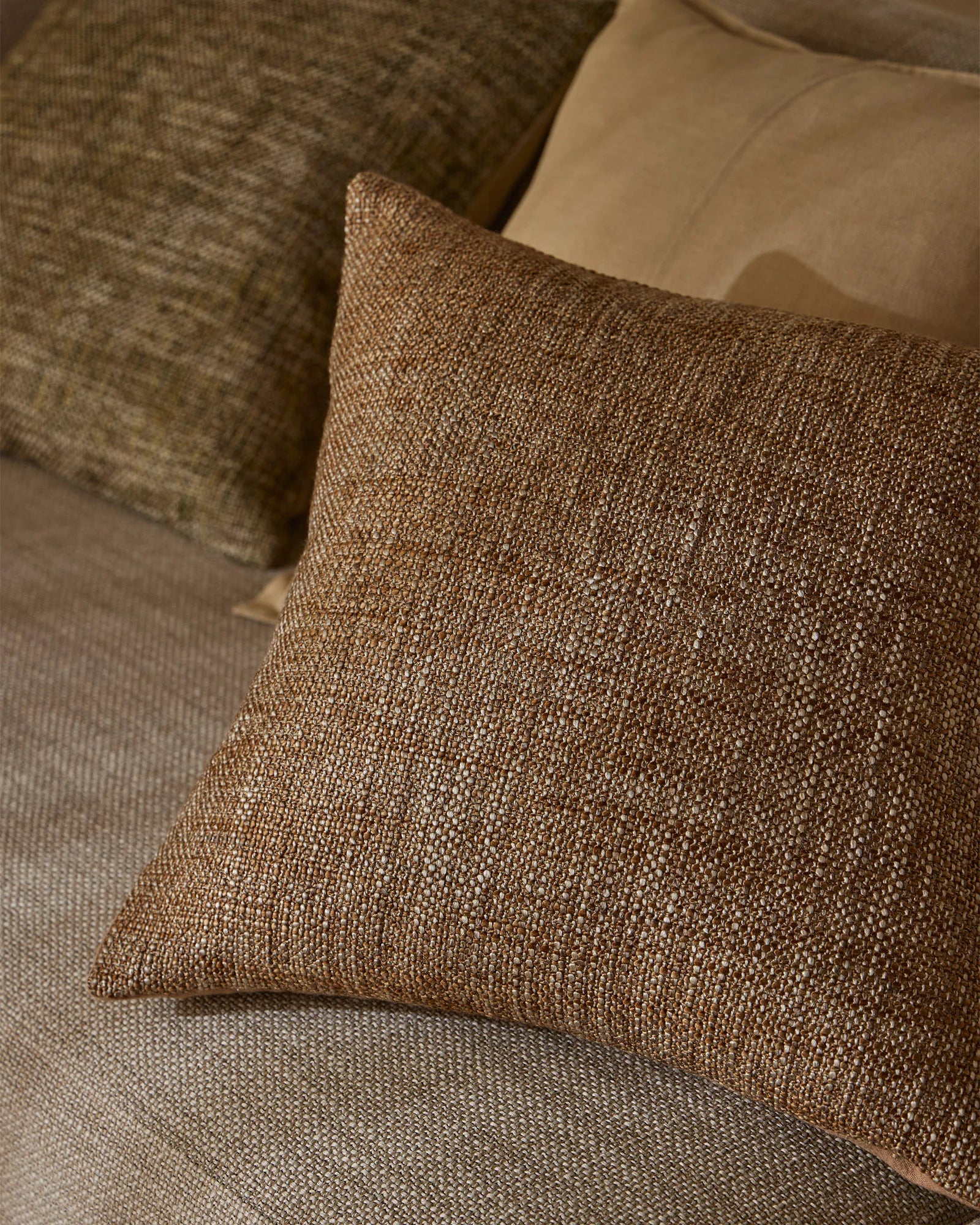 Warm neutral cushions in taupe and toffee tones layered on a cream upholstered sofa, showcasing textured woven fabrics and earthy minimalist interior styling