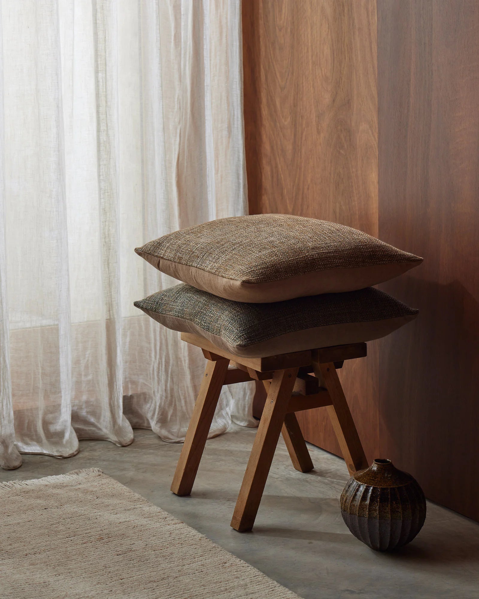 Stacked linen cushions in neutral tones on a wooden mid-century stool, styled with sheer cream curtains and warm wood panelling in a minimalist interior setting