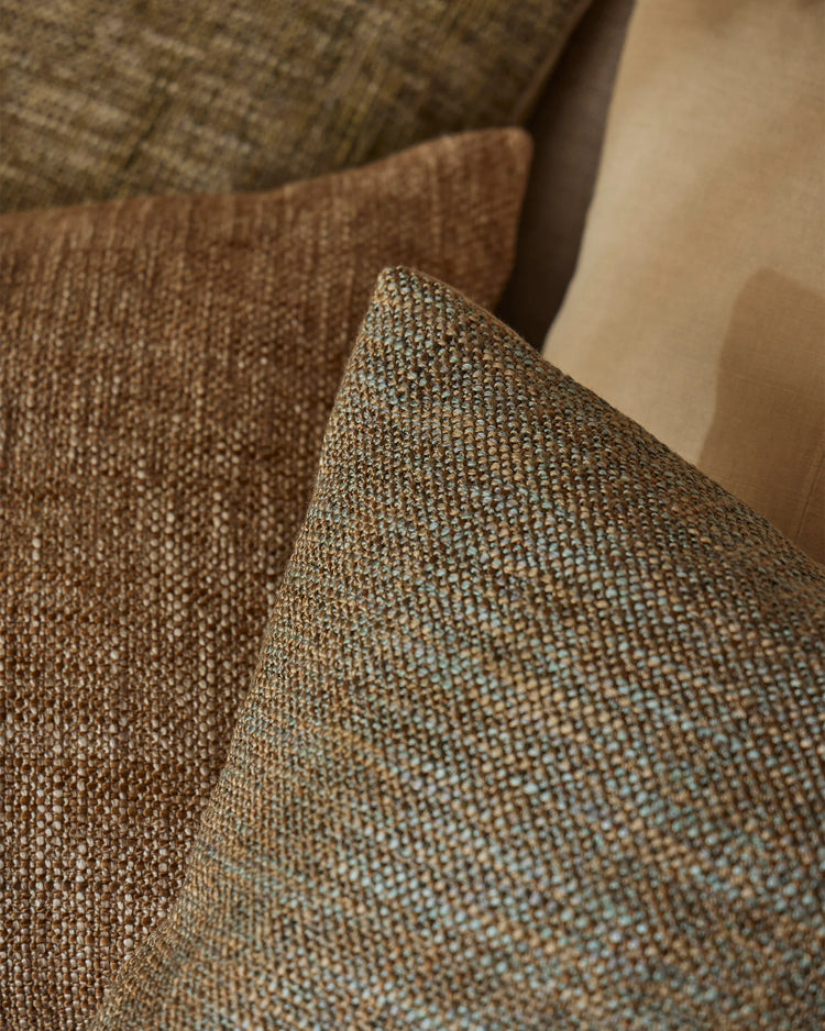 Earthy textured cushions in warm toffee and moss tones layered on neutral upholstery, highlighting woven fabric detail and warm minimalist home styling