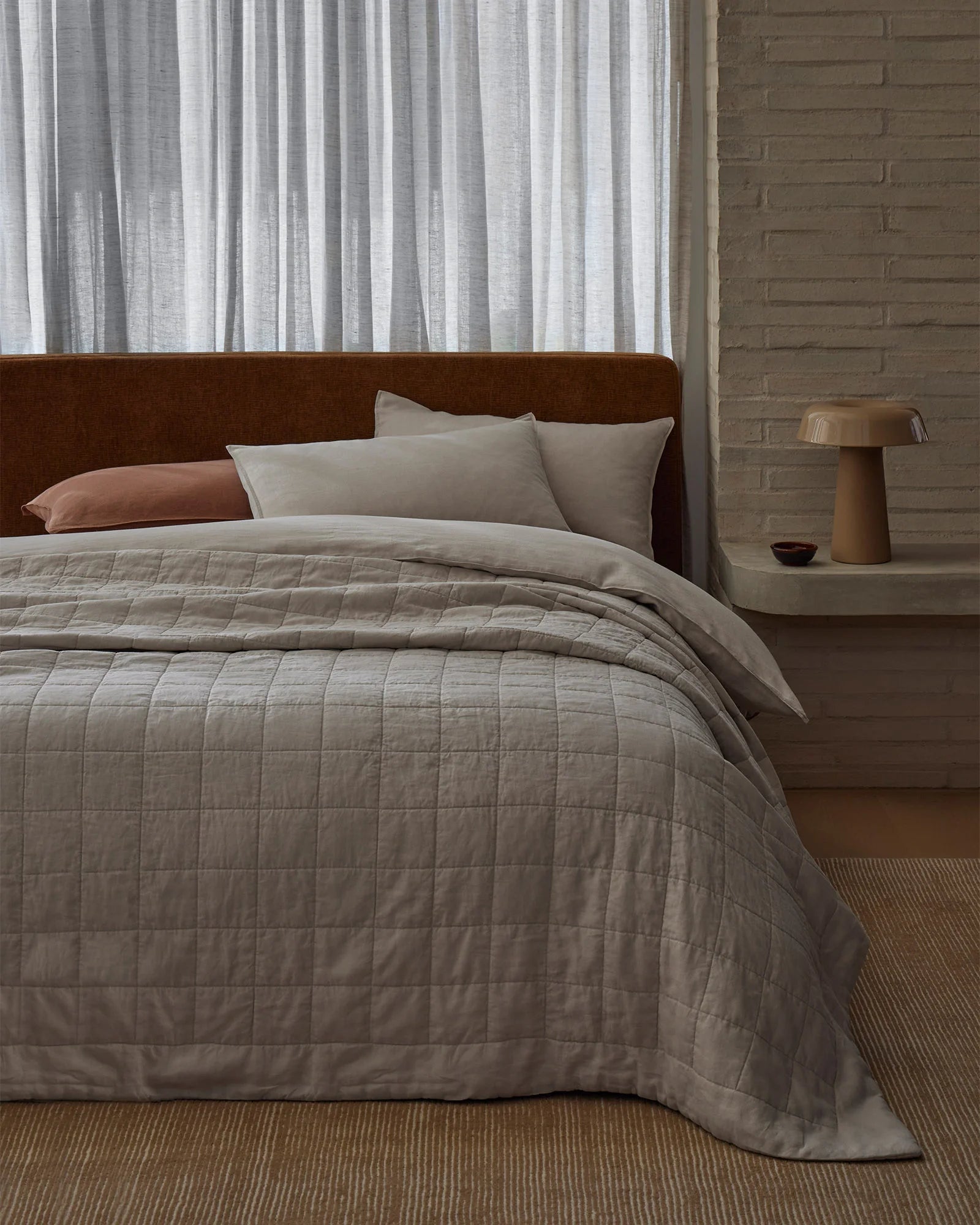 Minimalist bedroom with bone linen coverlet layered over neutral bedding, terracotta accent pillow, brown suede headboard, and sheer striped curtains in contemporary earthy palette
