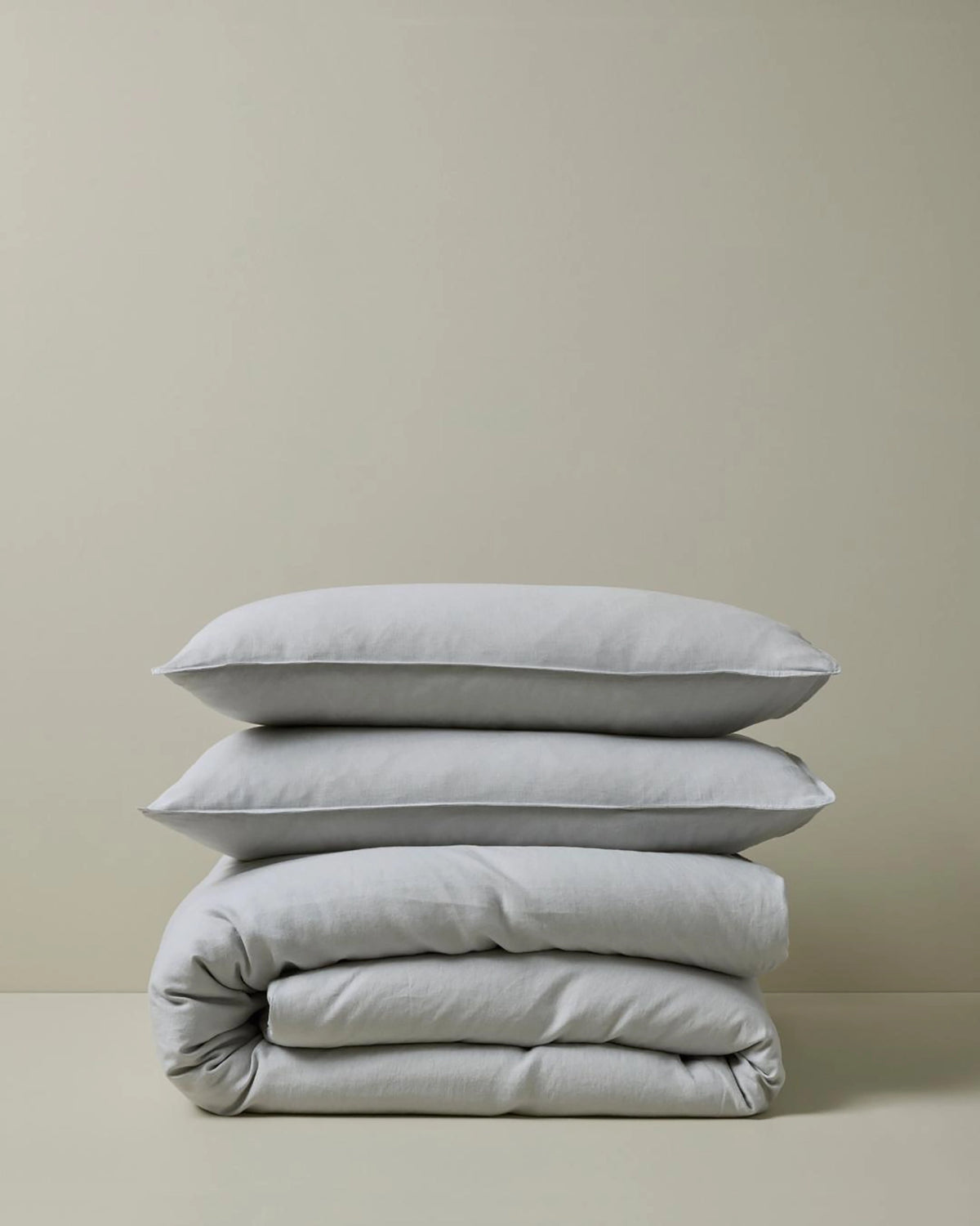 Stacked neutral linen bedding in soft silver tones, featuring Ravello quilt cover and pillowcases with natural texture and minimalist aesthetic against pale background