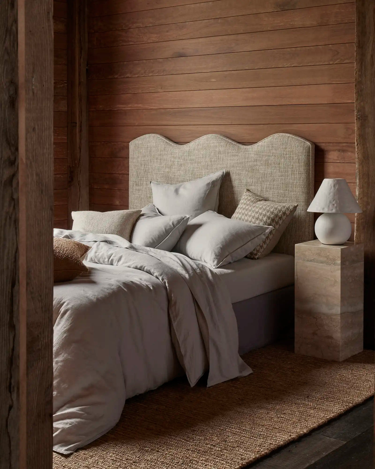 Neutral bedroom with dark timber walls and raw wood furnishings, styled with Ravello Quilt Cover in soft grey linen bedding layered with textured cushions and a knit throw