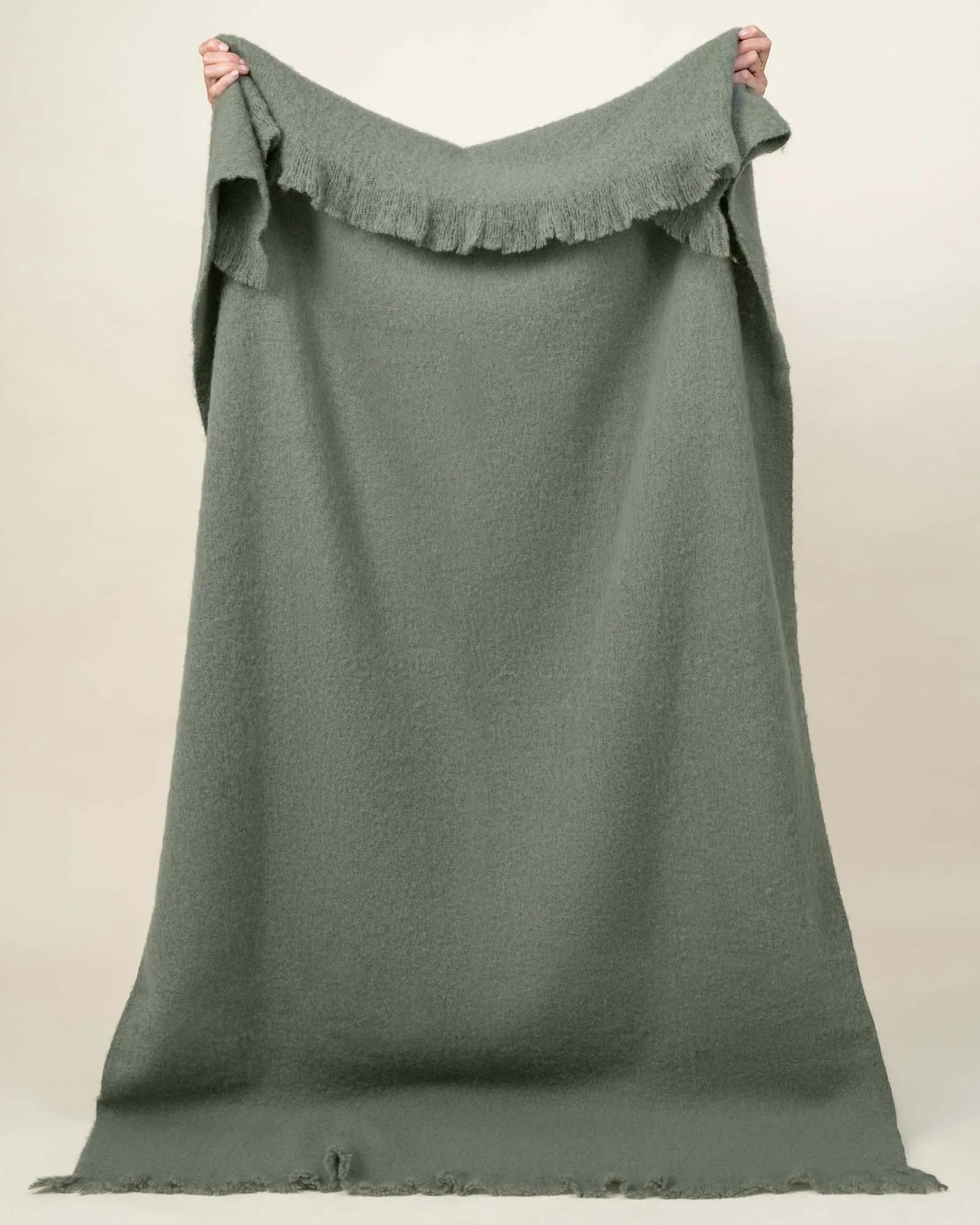 Hyde Oregano woven wool blend throw in sage green, held up to show fringed border detail by Weave Home
