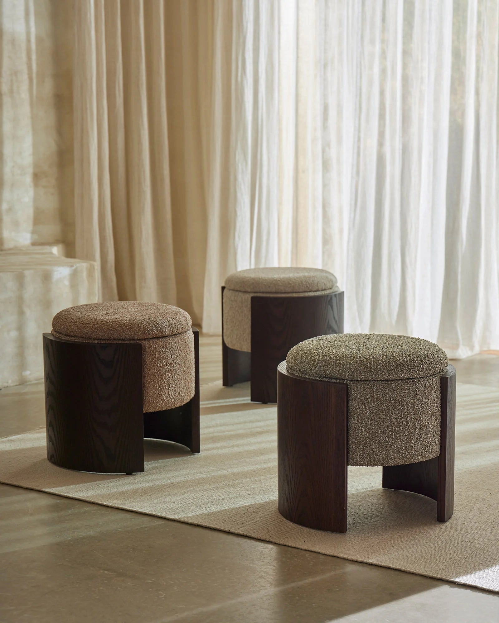 Three neutral-toned Norcia ottomans in moss, sienna and stone arranged on a cream area rug, featuring woven upholstery and dark timber bases, styled against floor-to-ceiling sheer curtains in a minimalist, earthy interior