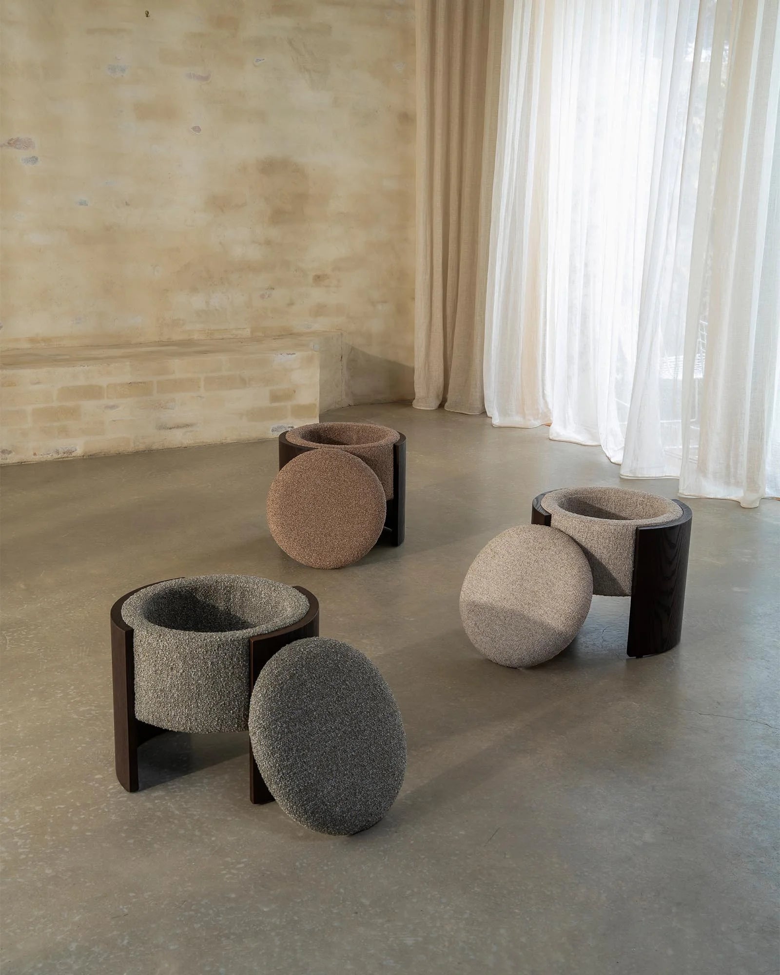 Norcia ottoman trio in earthy wool-blend tones—moss, sienna, and stone—arranged on polished concrete floor with sheer cream drapes and natural stone wall in minimalist interior