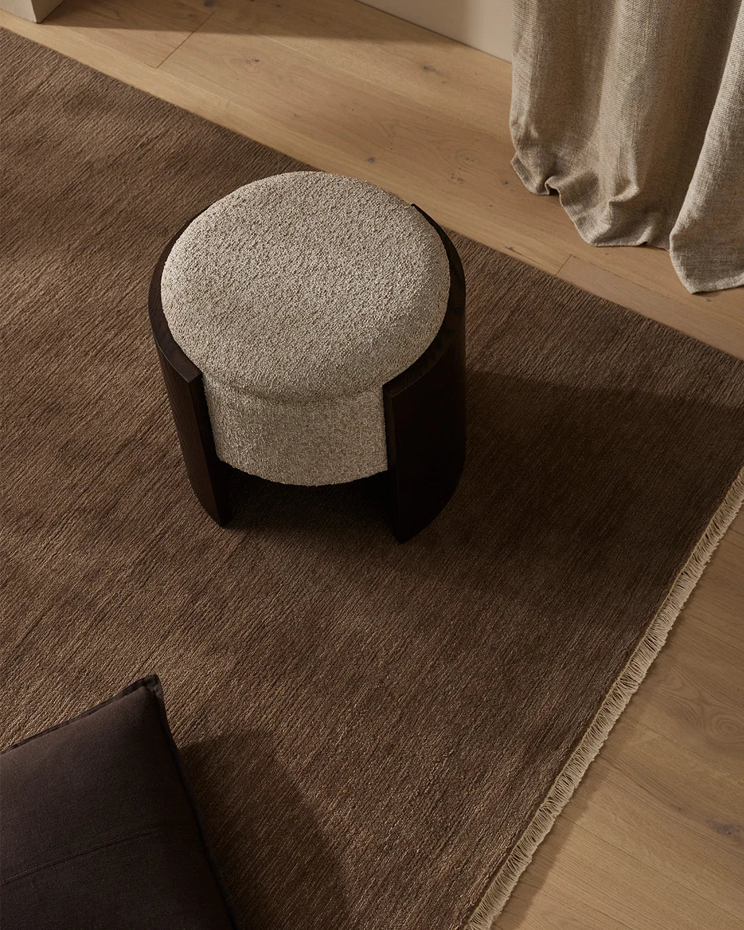 Neutral tonal living room with stone ottoman on textured taupe rug, warm timber flooring, linen curtains, and minimalist aesthetic