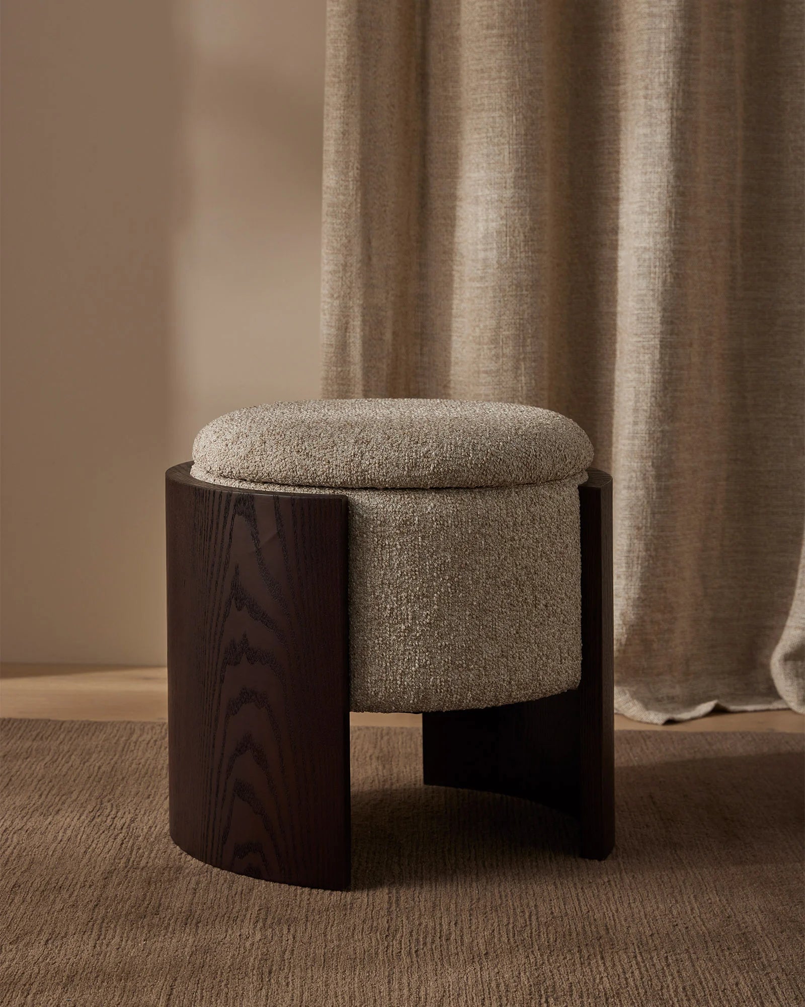 Warm neutral interior with taupe linen curtains, dark wood ottoman with oatmeal upholstered seat, and natural sisal rug creating a minimalist, earthy aesthetic.