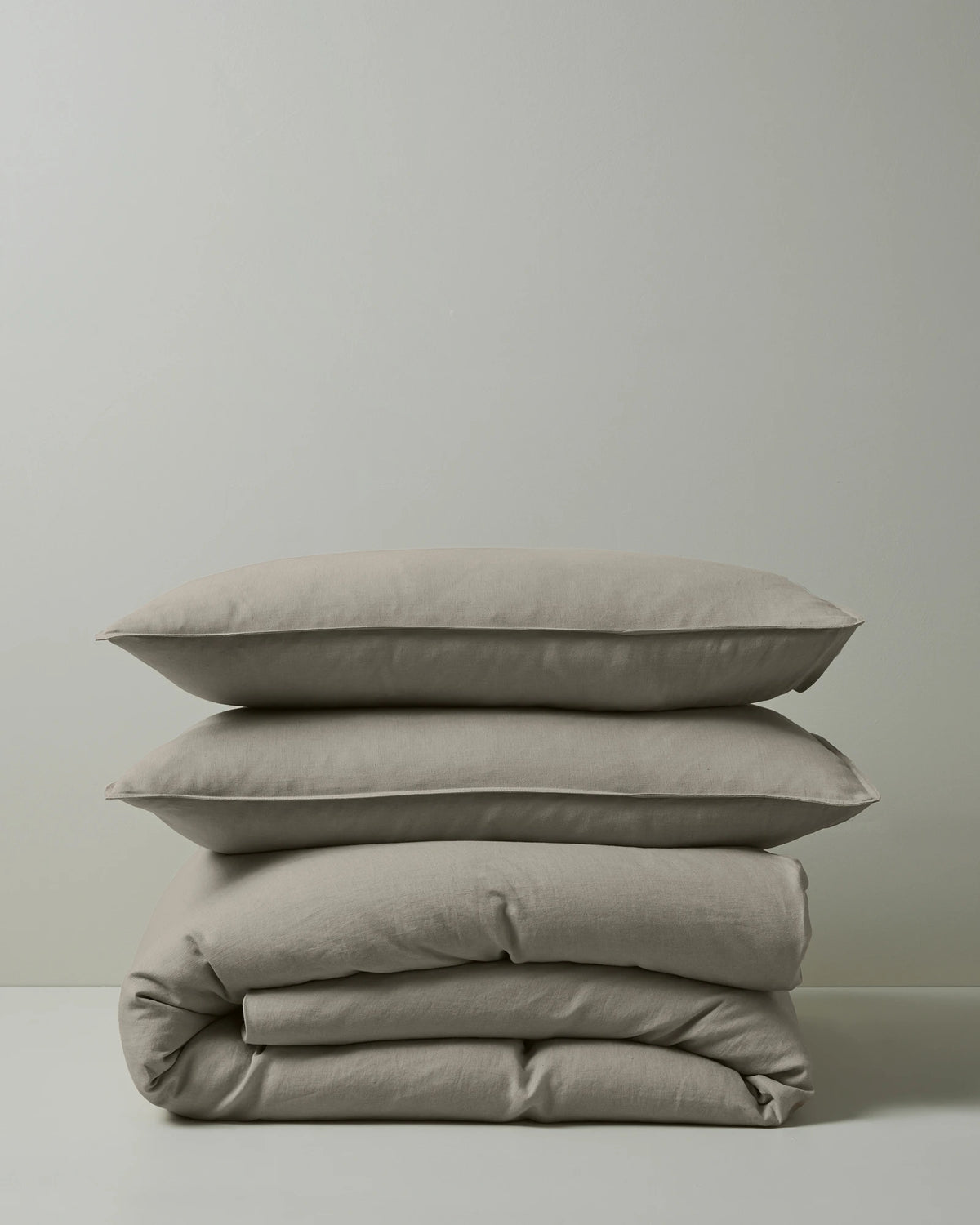 Stacked neutral linen bedding in soft pebble tones—Ravello pillowcases and quilt cover—showcasing minimalist styling with natural texture and understated elegance against a grey wall.