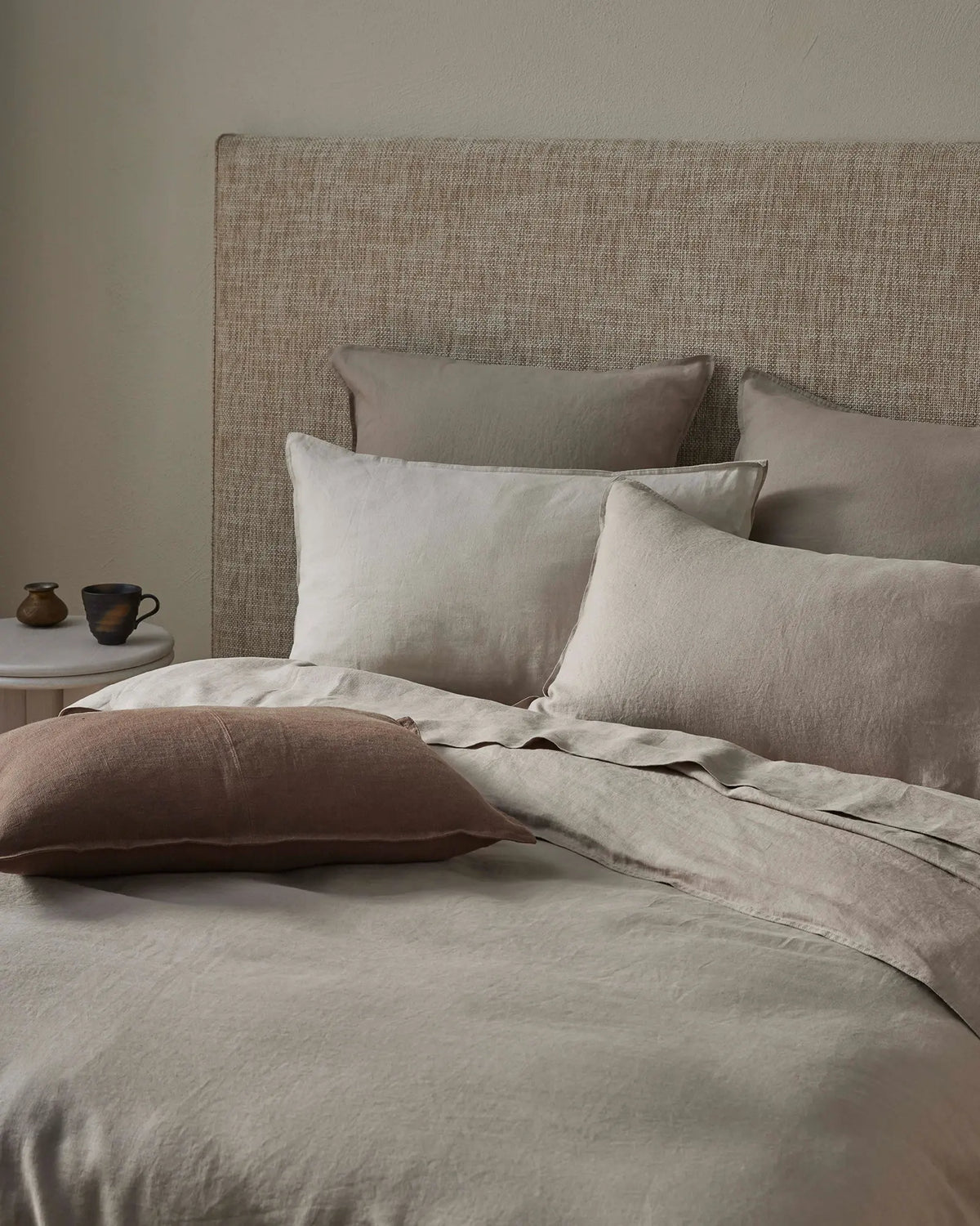 Neutral bedroom scene with layered linen bedding in soft pebble and mocha tones, textured headboard, and warm minimalist styling