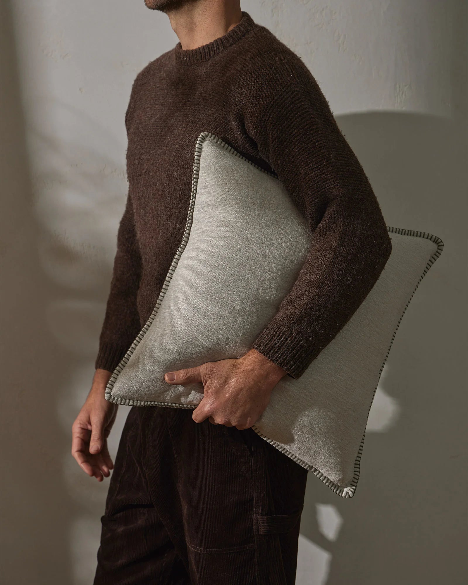 Warm-toned lifestyle scene: man holding Alba cushion in bone with contrast whipstitched edge, layered with chocolate knit sweater and charcoal corduroy against neutral wall backdrop